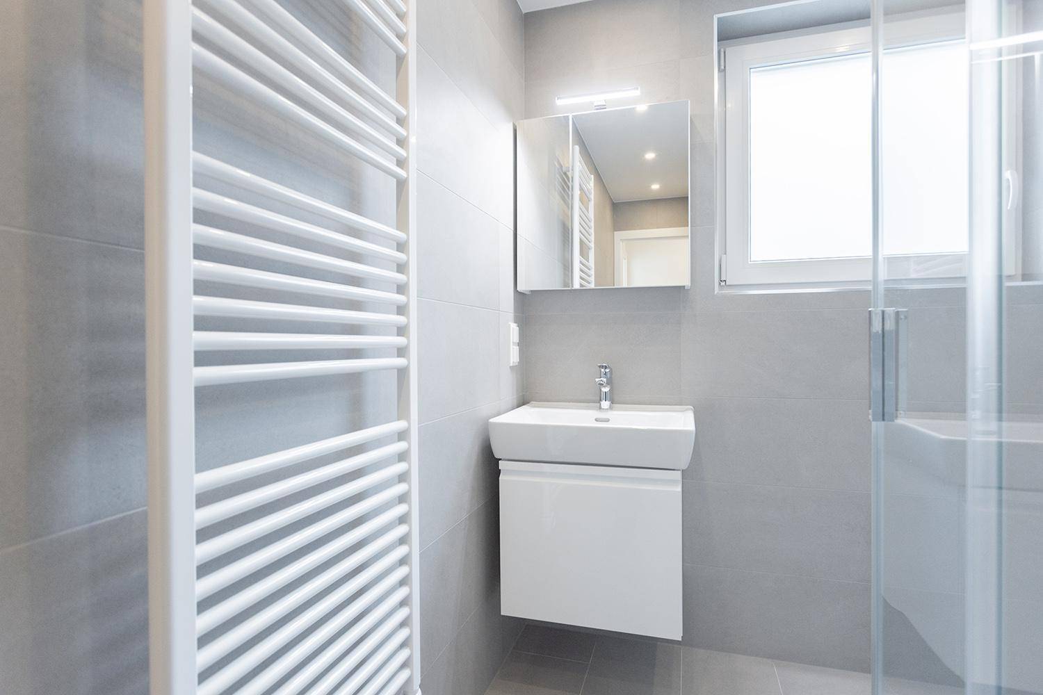 Modernes Badezimmer mit Dusche, Waschbecken, Fenster und beheiztem Handtuchhalter in hellen Grautönen.