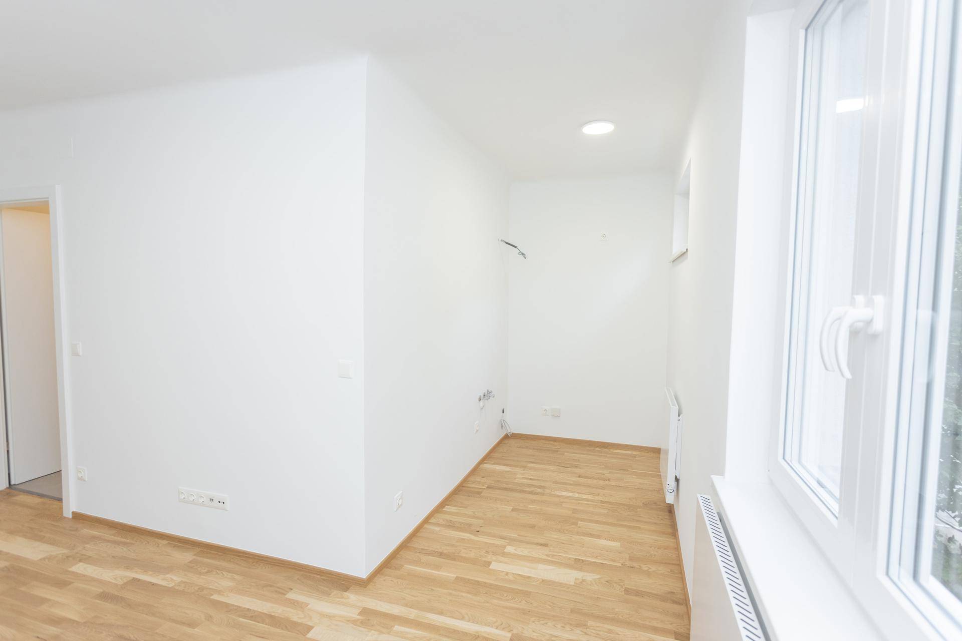 Schmaler, heller Raum mit Holzboden, weiß gestrichenen Wänden und einem Fenster.