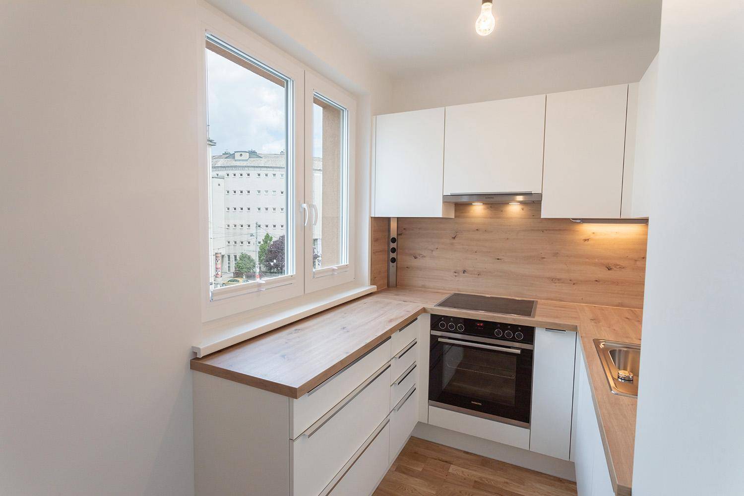 Helle Küche mit Fenster, weißen Schränken und Holzarbeitsplatte, Blick auf die Stadt, funktional und hell.