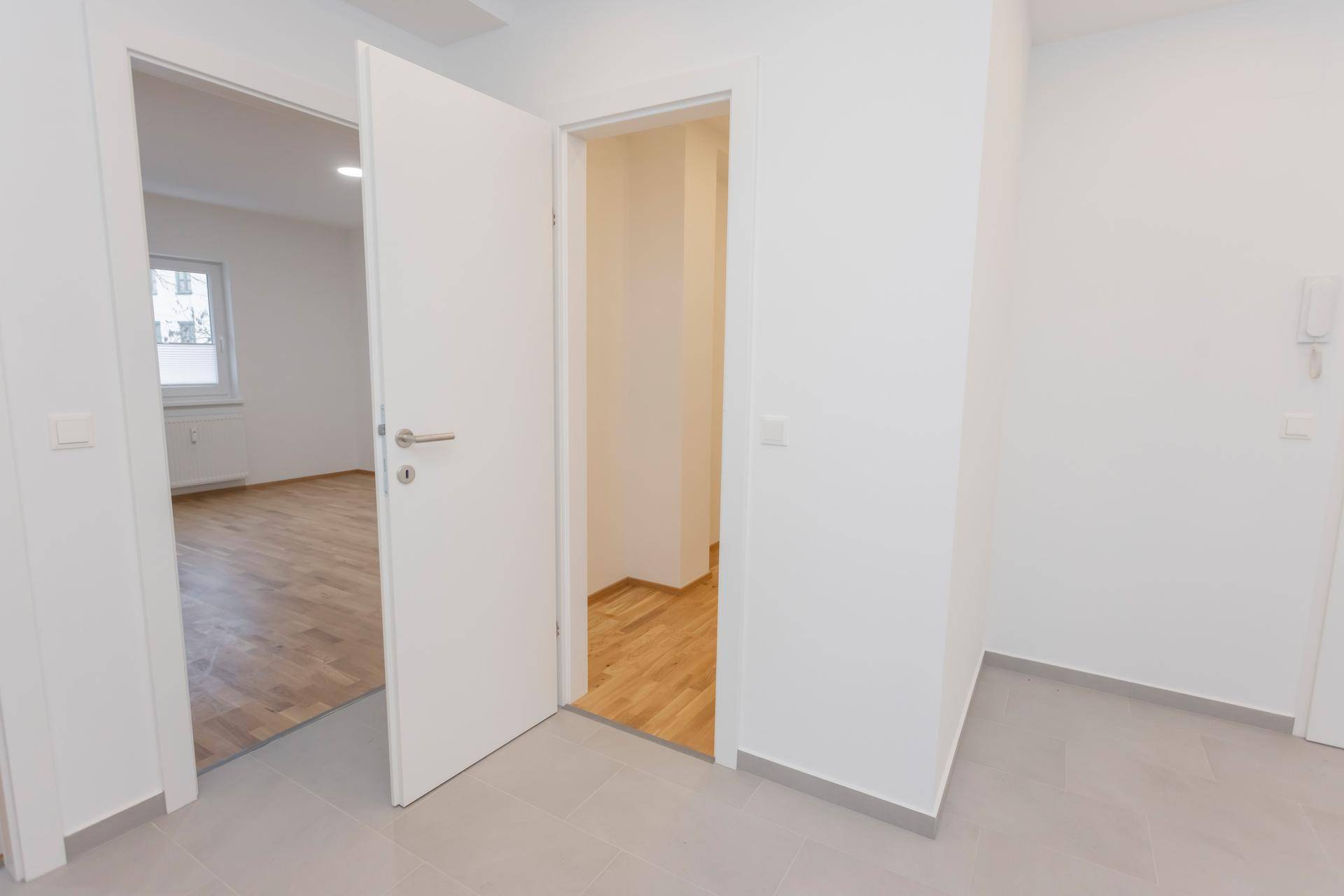 Heller Flur mit grauen Fliesen und offener Tür zu einem Zimmer mit Parkettboden.
