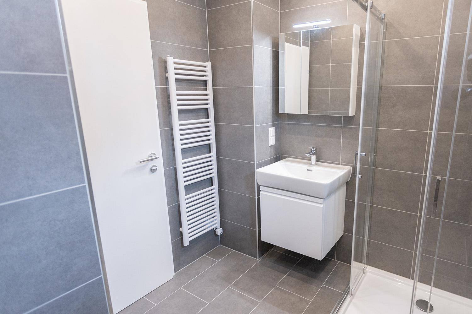 Modernes Badezimmer mit grauen Fliesen, Handtuchheizkörper und Waschtisch, hell und gepflegt.