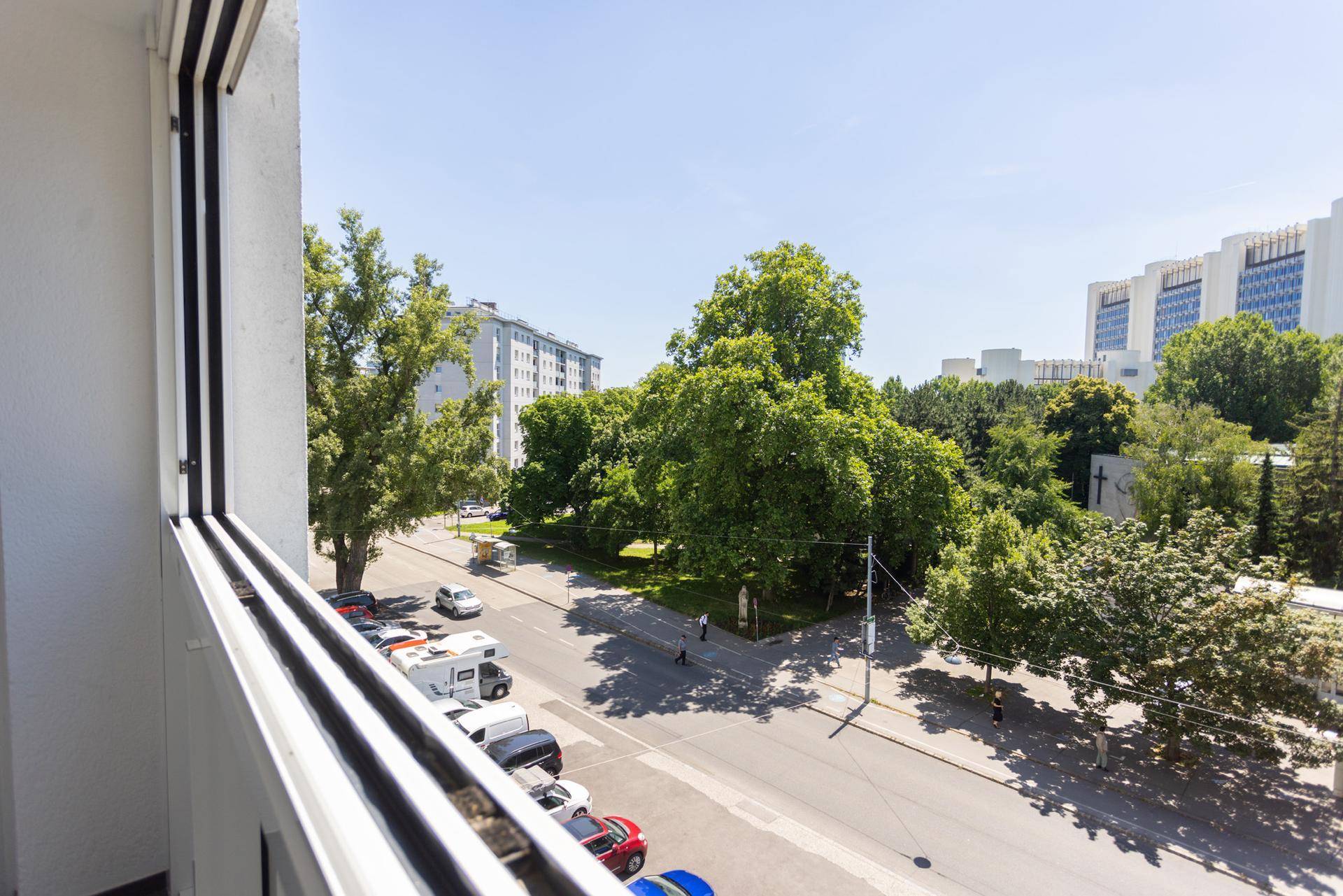 Weitblick über eine belebte Straße mit Bäumen und umliegenden Gebäuden unter blauem Himmel.
