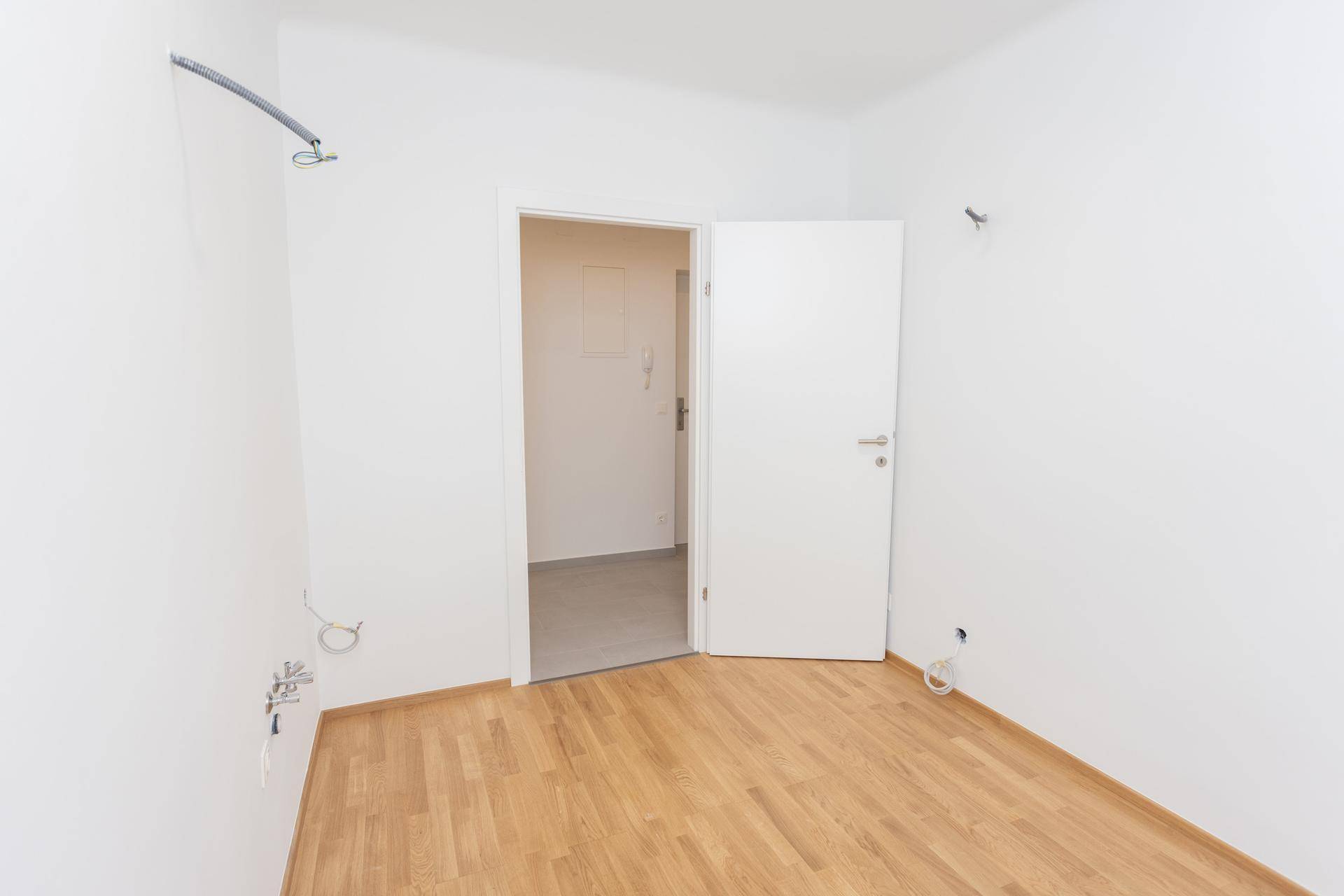 Leeres Zimmer mit Holzparkettboden und offener Tür, die den Blick in den Flur freigibt.