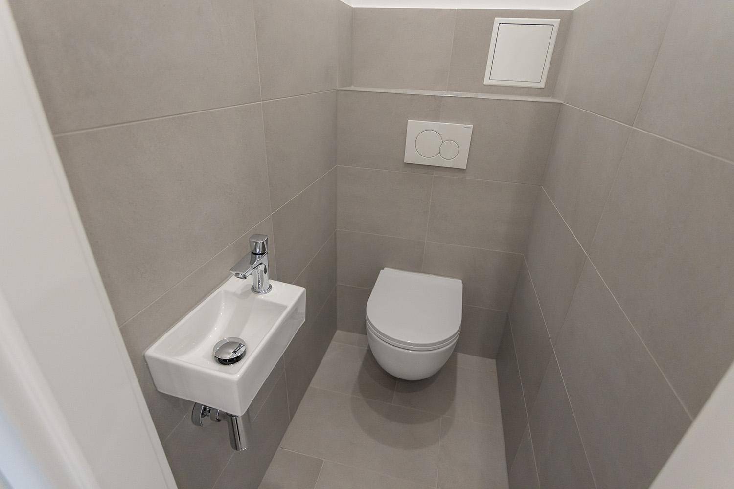 Separate, moderne Gästetoilette mit grauen Fliesen, wandhängendem WC und kleinem Handwaschbecken.