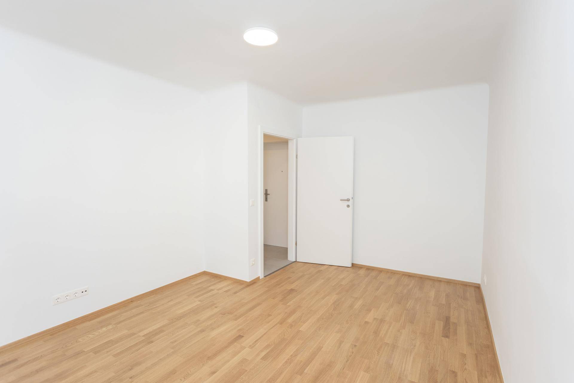 Leeres, helles Zimmer mit Holzparkettboden und einer Tür, die zu einem anderen Raum führt.