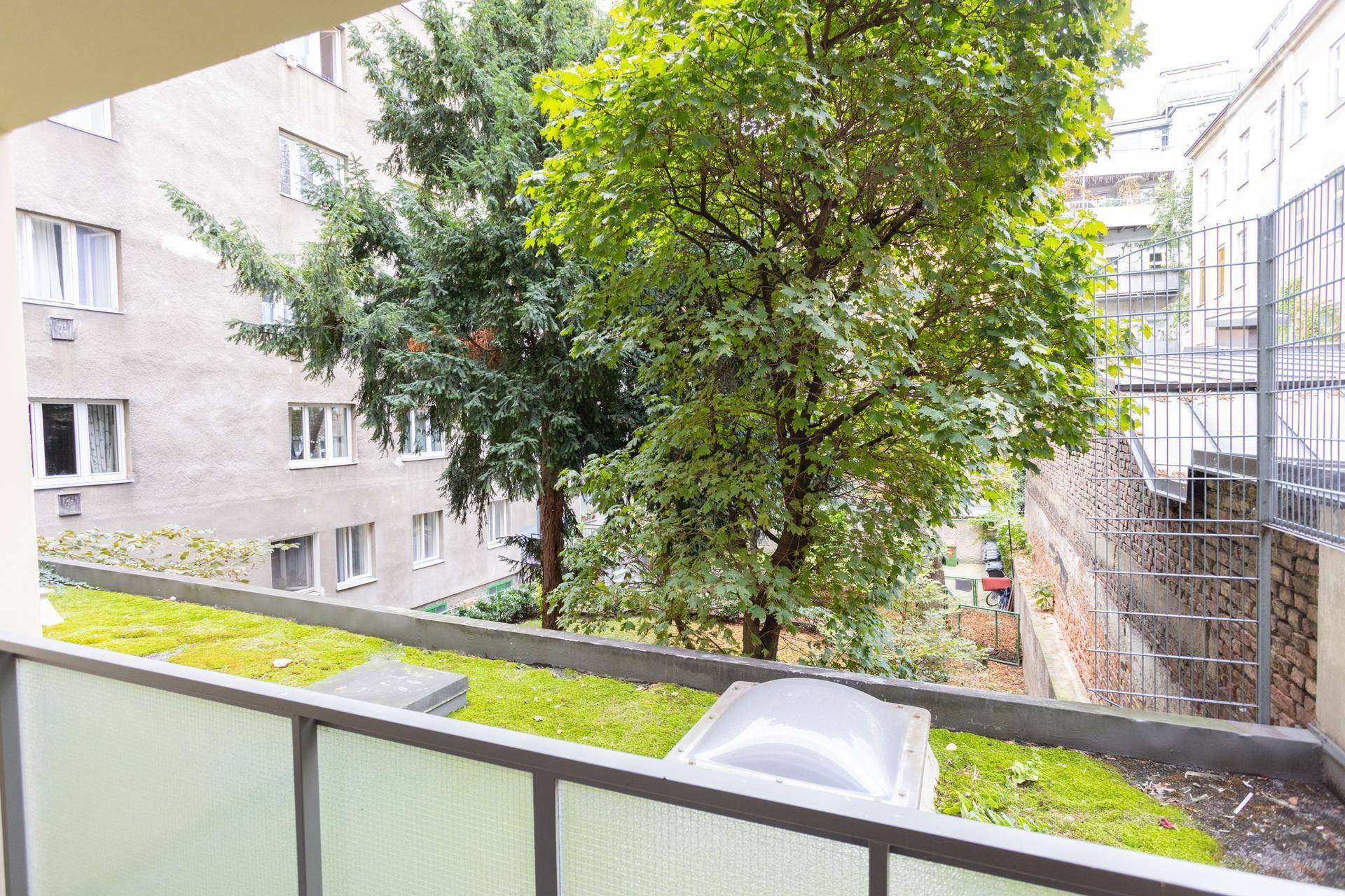Blick von einem Balkon auf üppige Bäume, eine begrünte Fläche und umliegende Gebäude.
