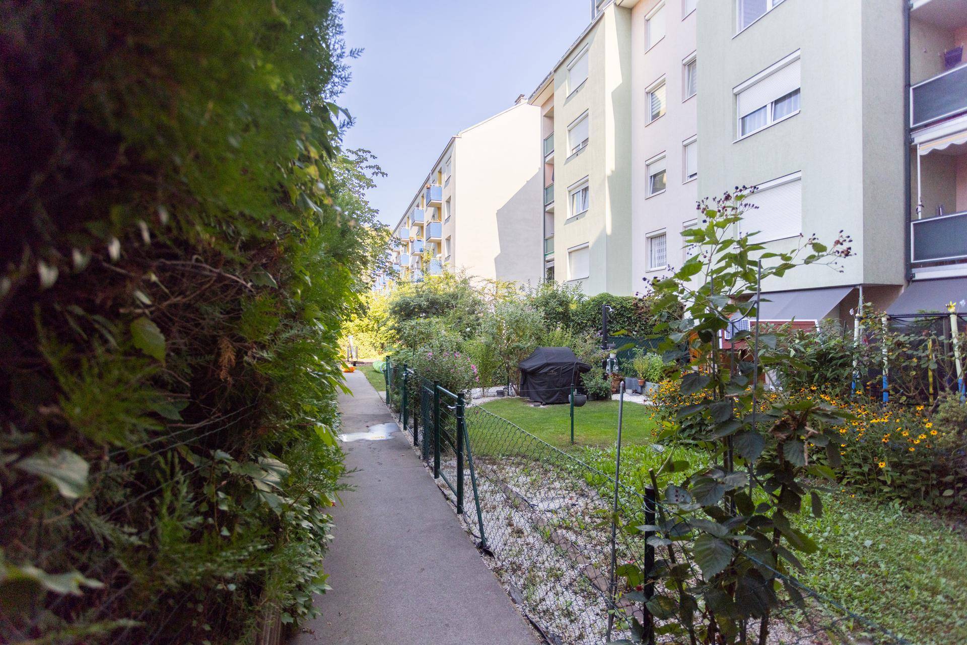 Grüner Gemeinschaftsgarten mit Hecken, Bäumen und einem Weg, umgeben von Wohngebäuden.