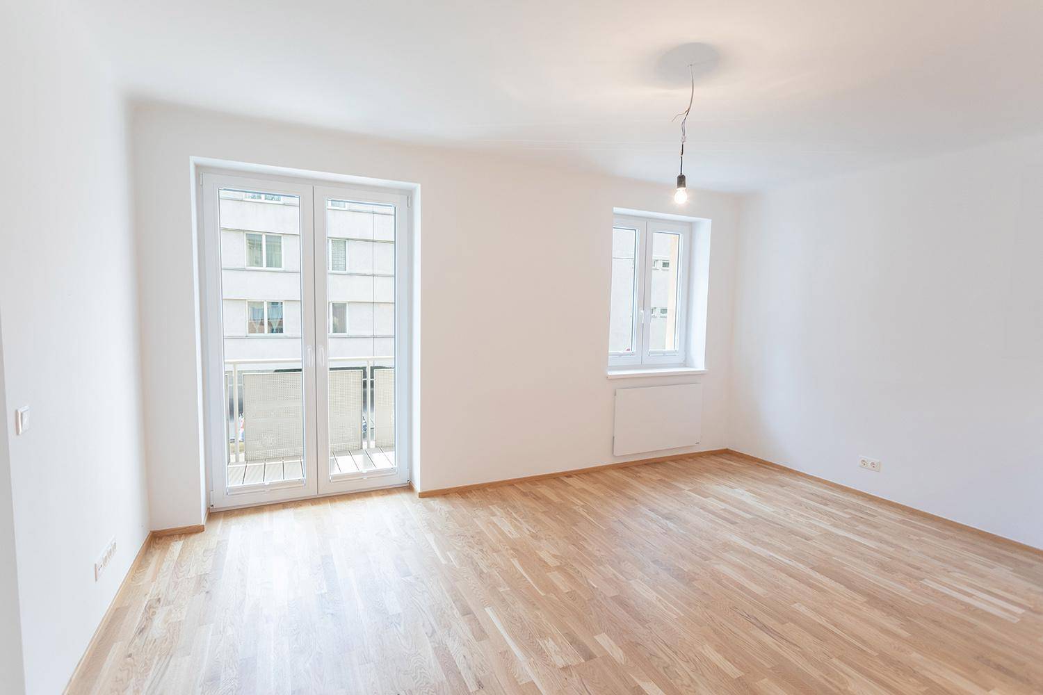 Helles, leeres Zimmer mit Parkettboden und Zugang zu einem Balkon.