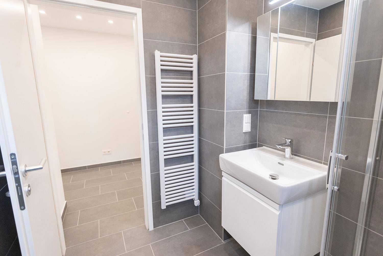Modernes Badezimmer mit grauen Fliesen, Handtuchheizkörper und Waschtisch, mit Zugang zum Flur.