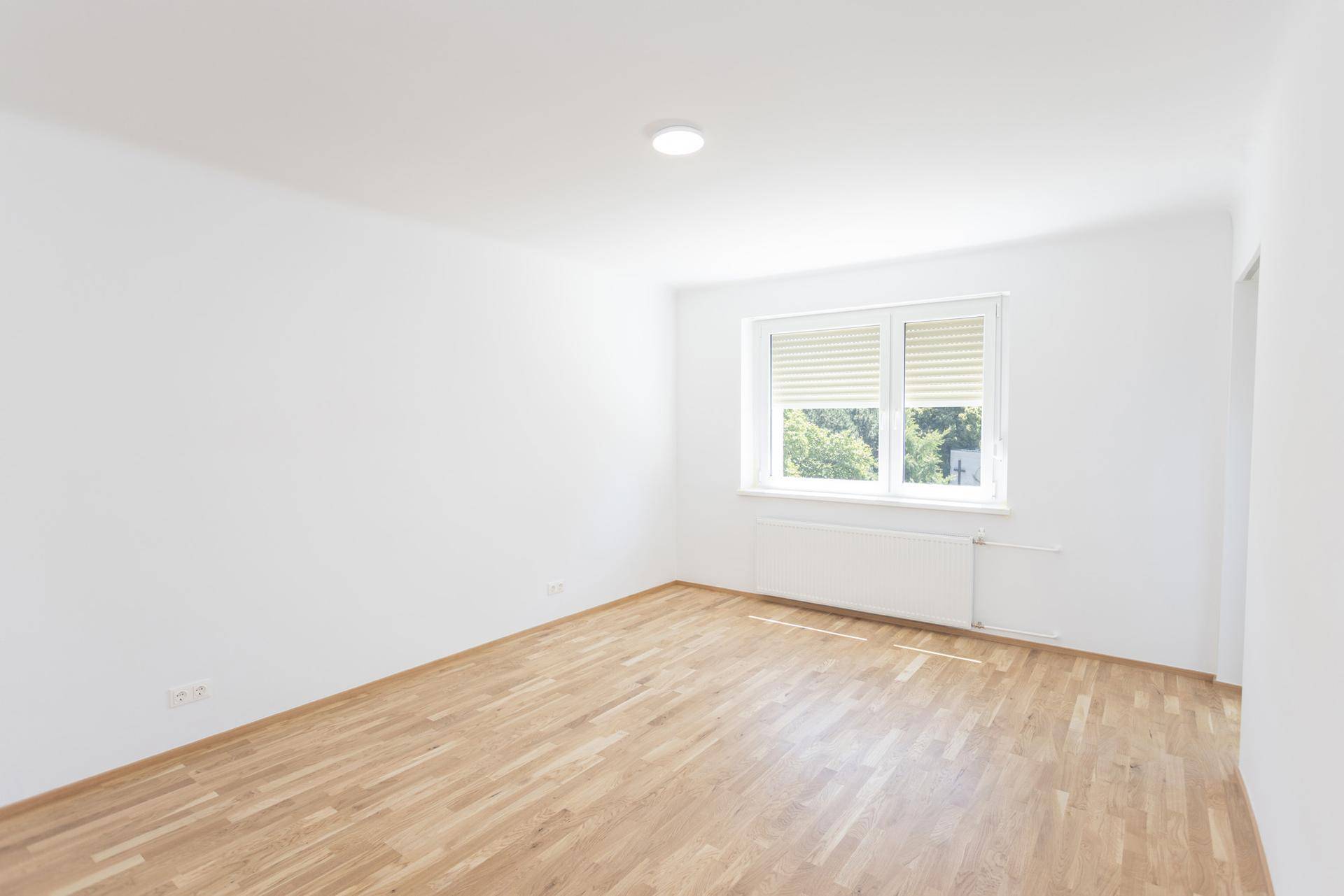 Helles, leeres Schlafzimmer mit Holzboden und Fenster, bereit zur Einrichtung.