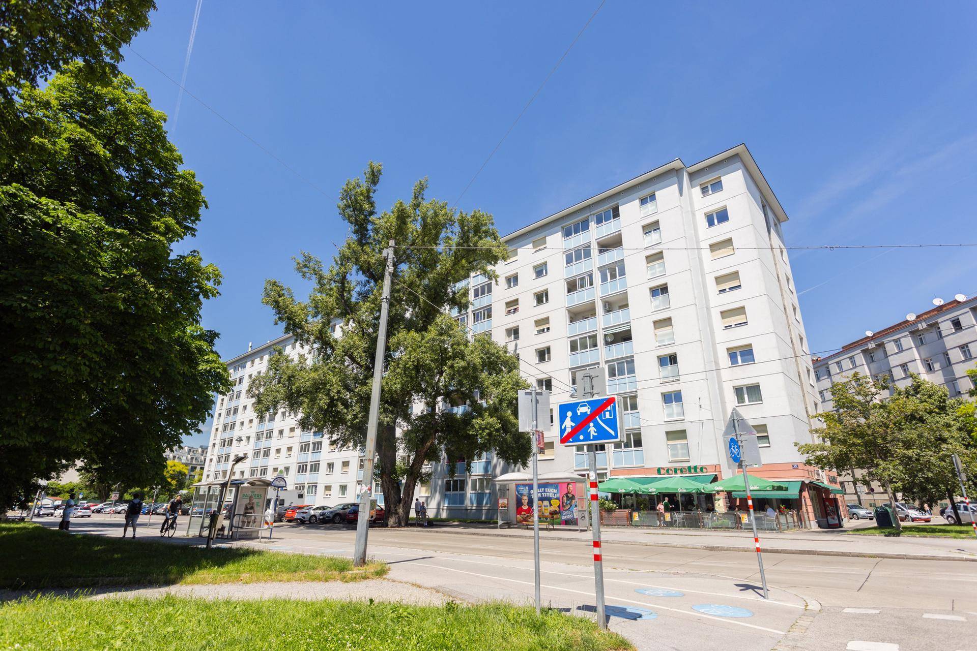 Mehrfamilienhaus an einer Straße mit Geschäften und viel Grün in der Umgebung.