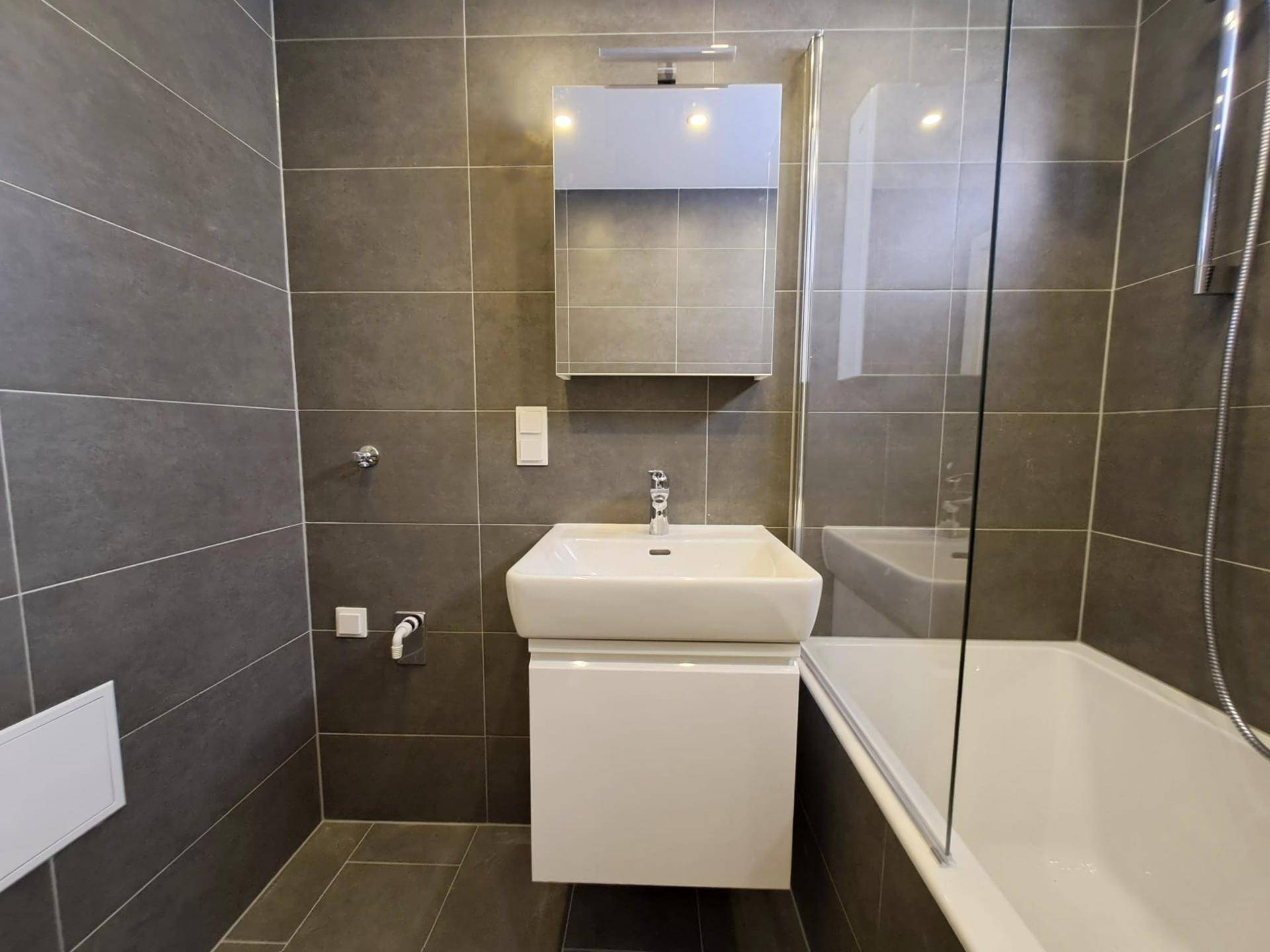 Modernes Badezimmer mit dunklen Fliesen, Waschbecken und Badewanne mit Duschabtrennung aus Glas.