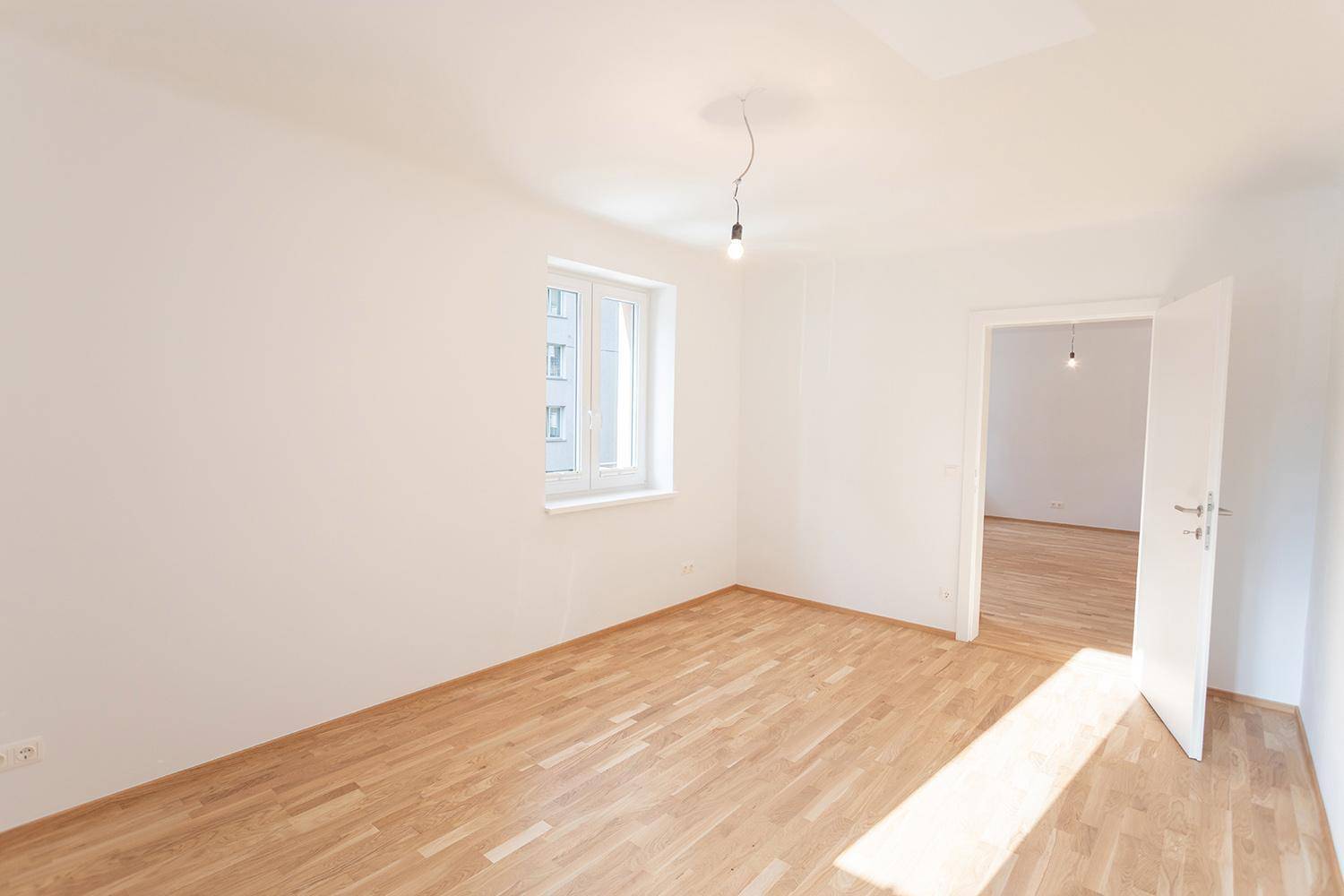 Helles Zimmer mit Parkettboden und Zugang zu einem weiteren, leeren Raum.