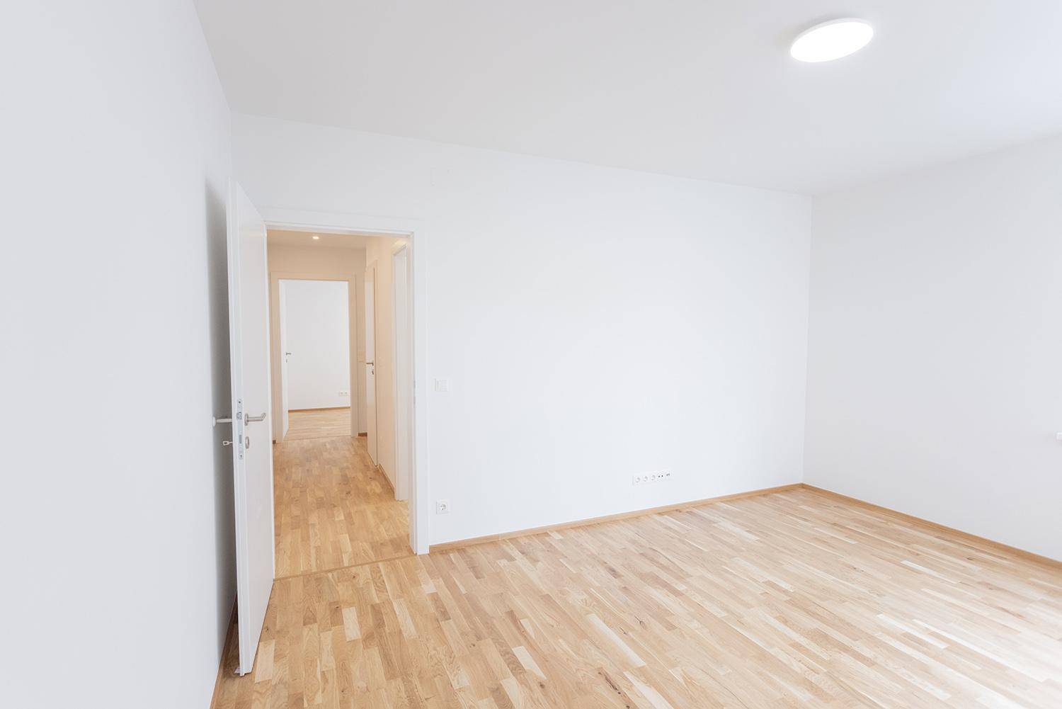 Leerer Raum mit Holzparkettboden und weiß gestrichenen Wänden, mit Blick in den Flur.