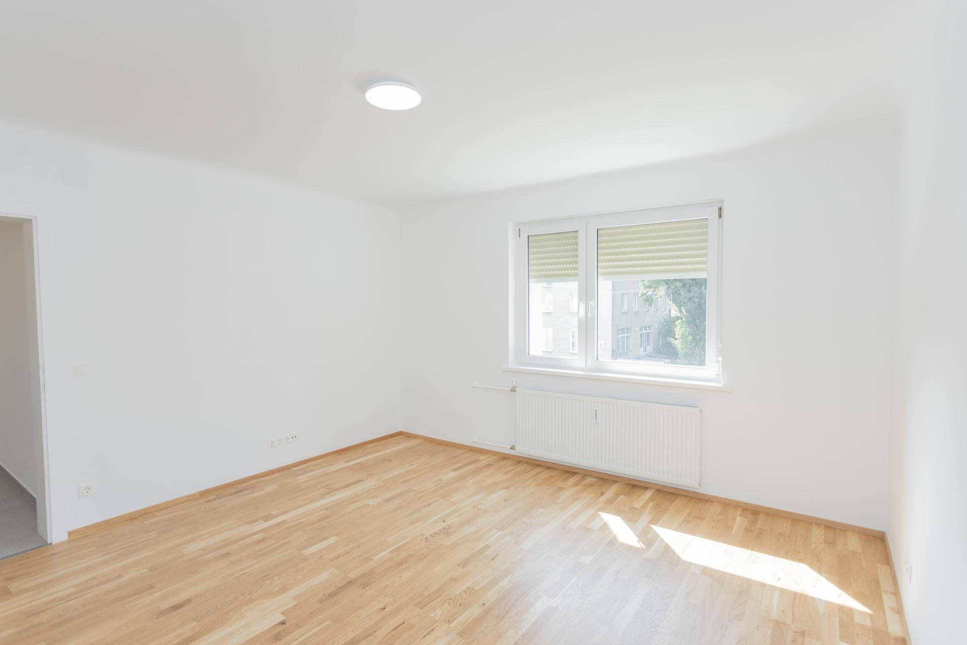 Leeres, helles Schlafzimmer mit Holzboden und Fenster, das einen Blick auf die Umgebung bietet.
