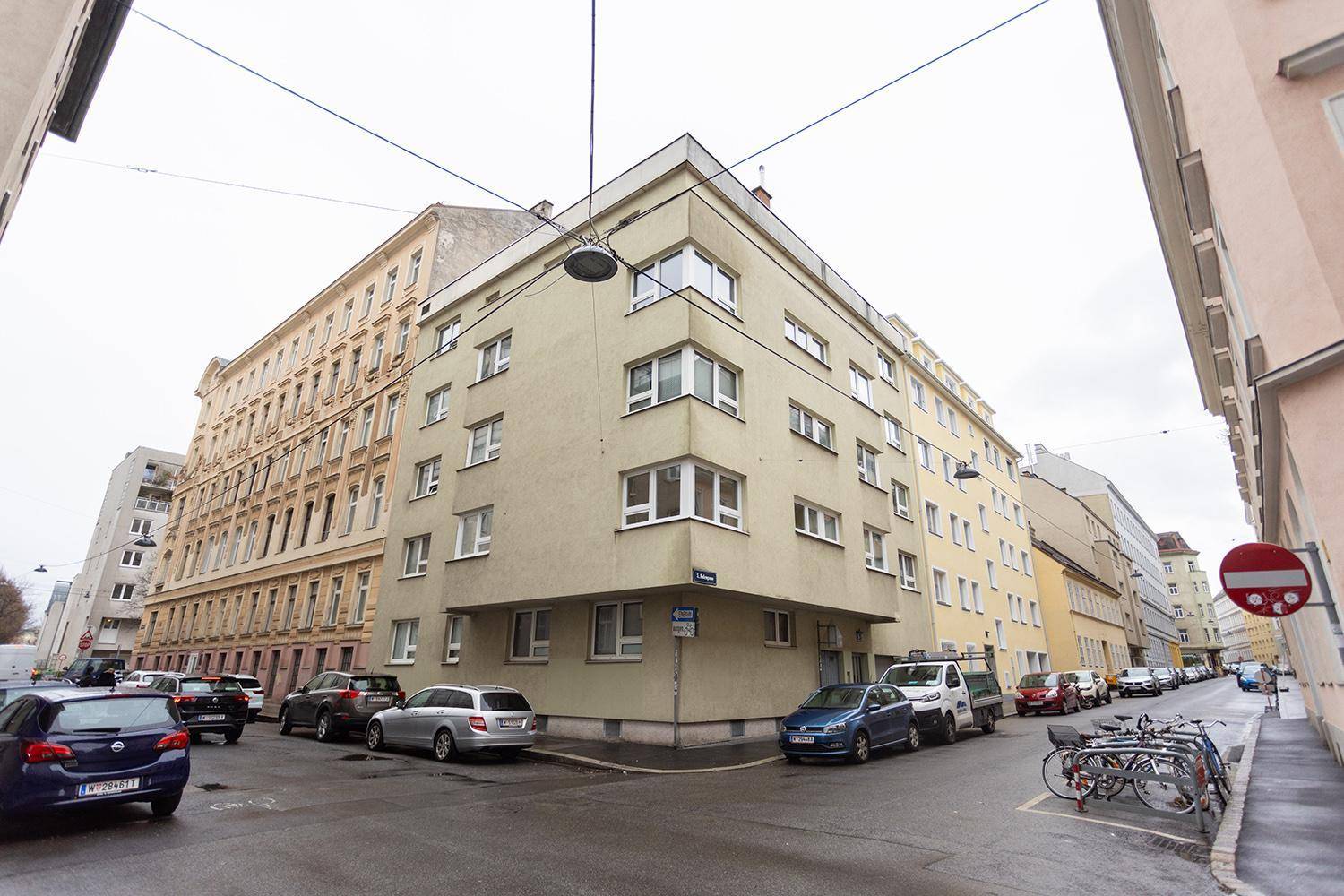 Eckgebäude in städtischer Umgebung mit mehreren Stockwerken und geparkten Autos auf der Straße.