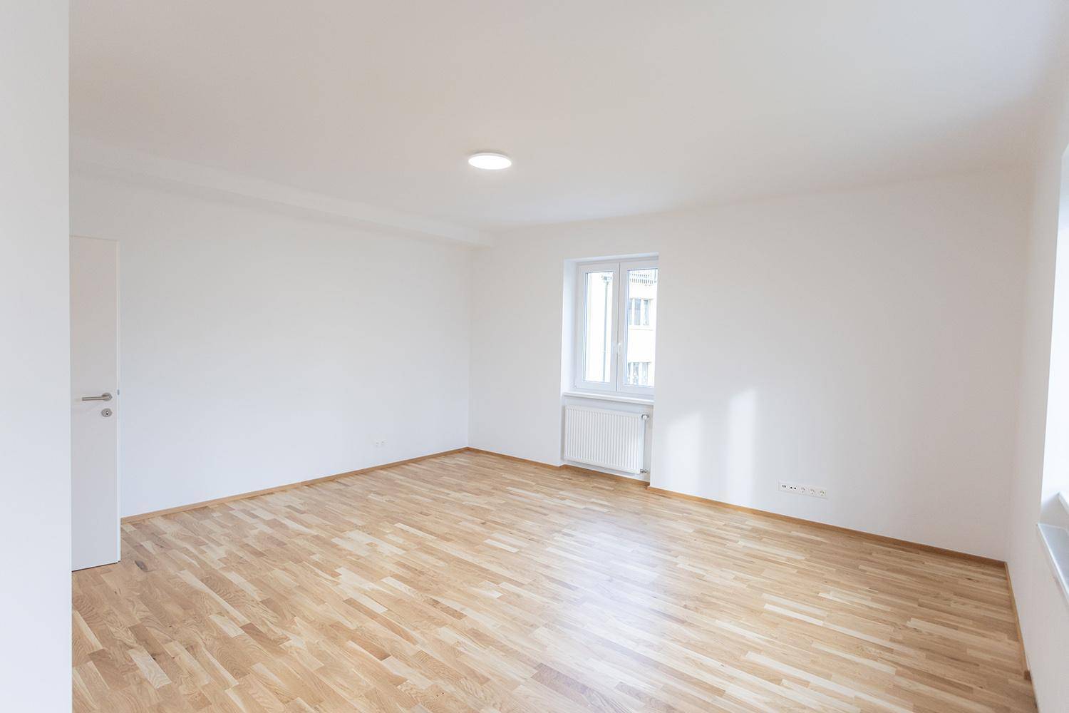 Helles Wohnzimmer mit Holzparkett, weißen Wänden und großem Fenster, unmöbliert.