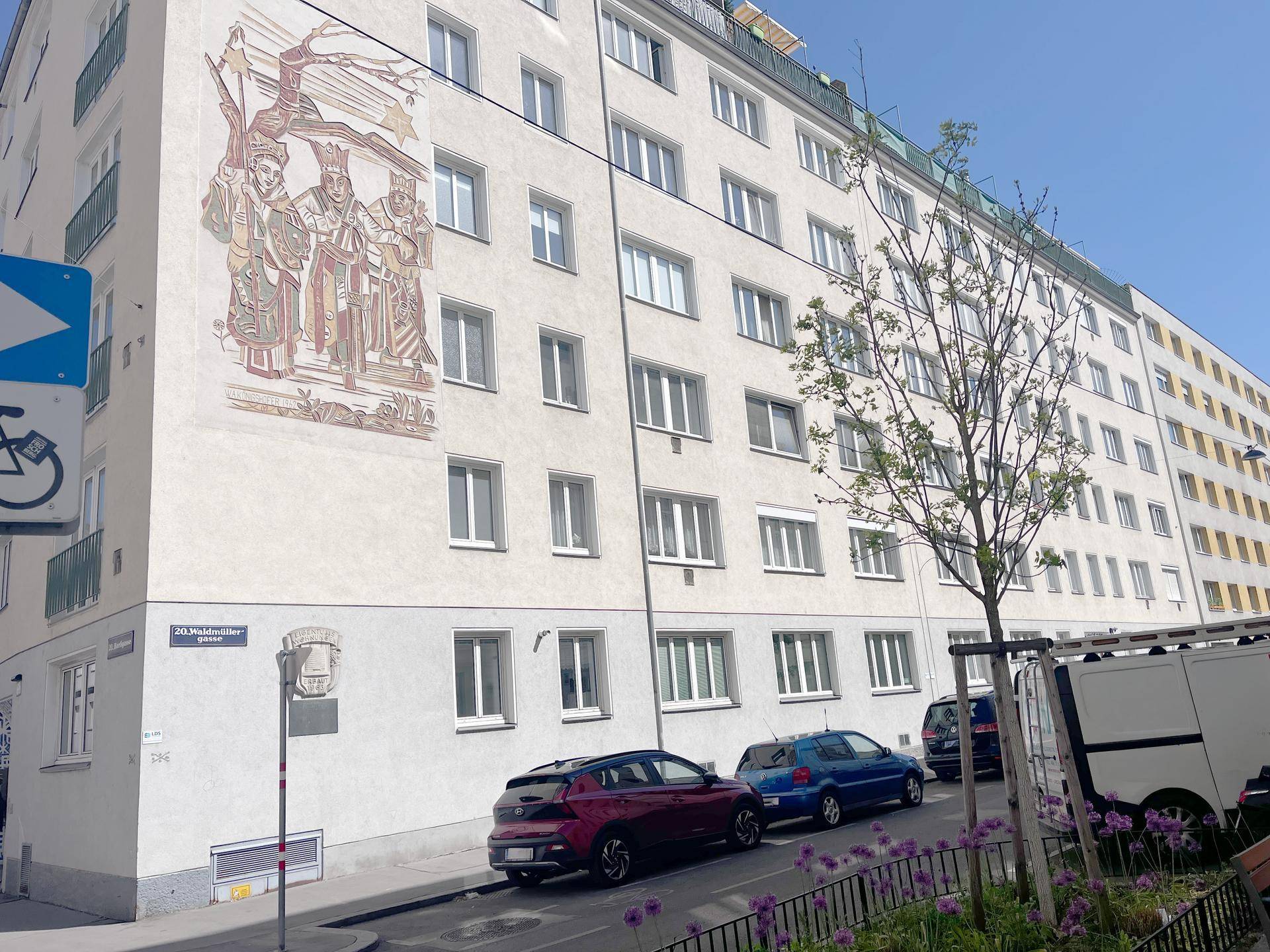 Wohngebäude mit Fassadenmalerei und parkenden Autos an einem sonnigen Tag.