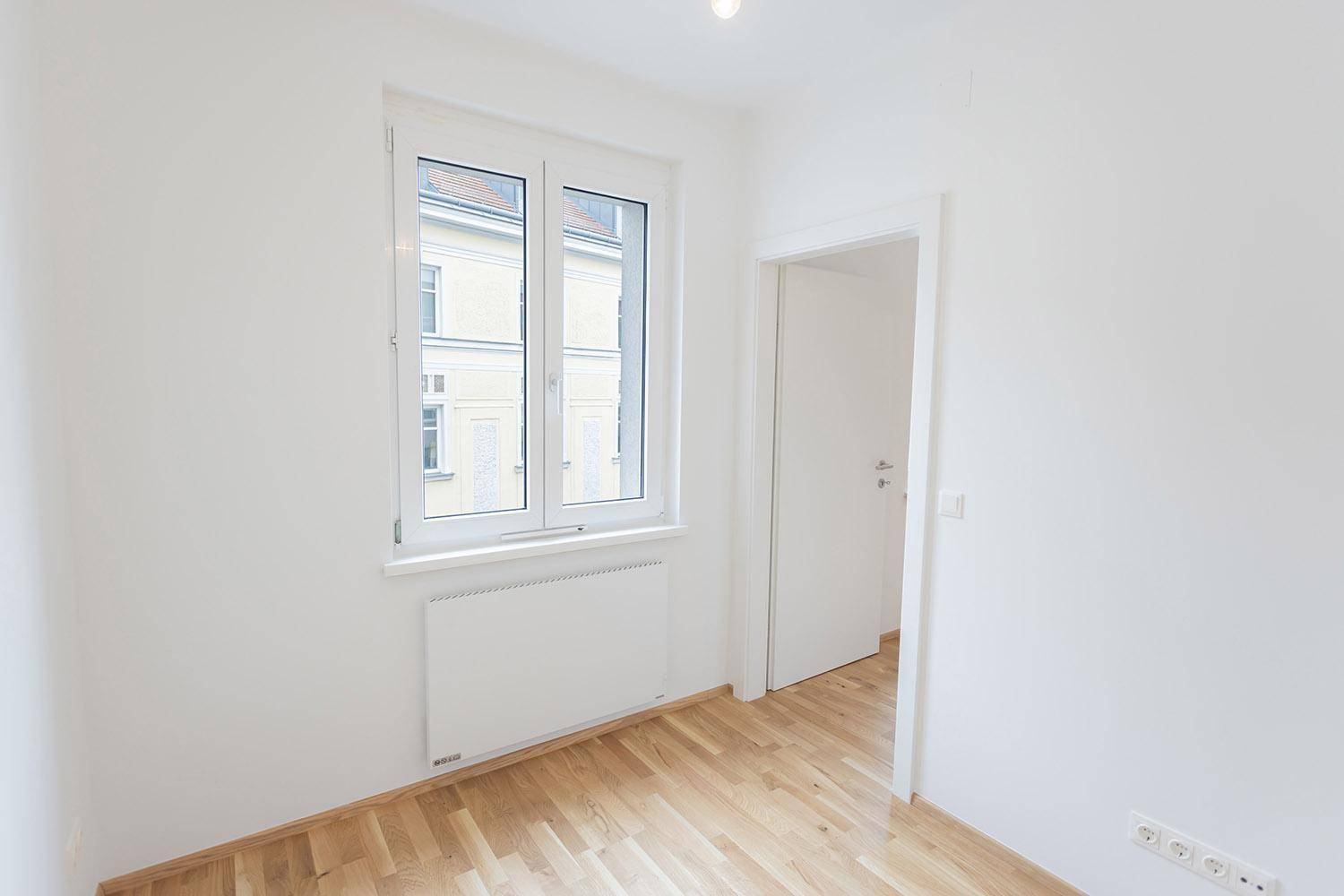 Helles, leeres Zimmer mit Parkettboden und einem Fenster, das auf ein gegenüberliegendes Gebäude blickt.