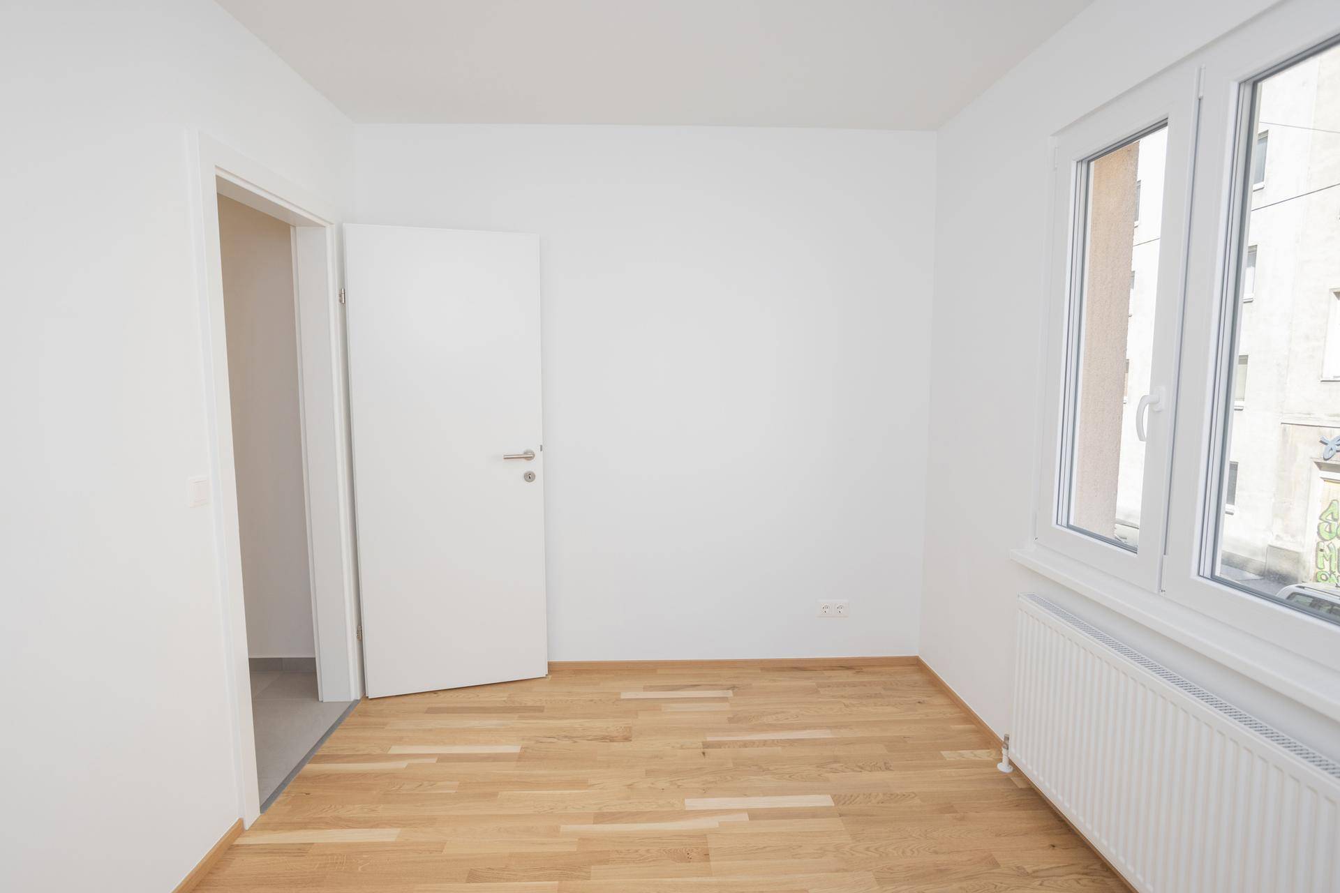 Unmöbliertes Zimmer mit hellem Parkettboden, weißen Wänden und einem Fenster mit Blick nach draußen.