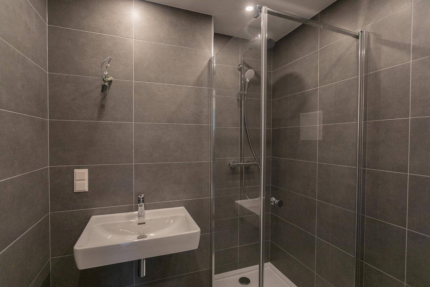 Zeitgemäßes Badezimmer mit Duschkabine, grauen Fliesen und Heizkörper als Handtuchhalter.