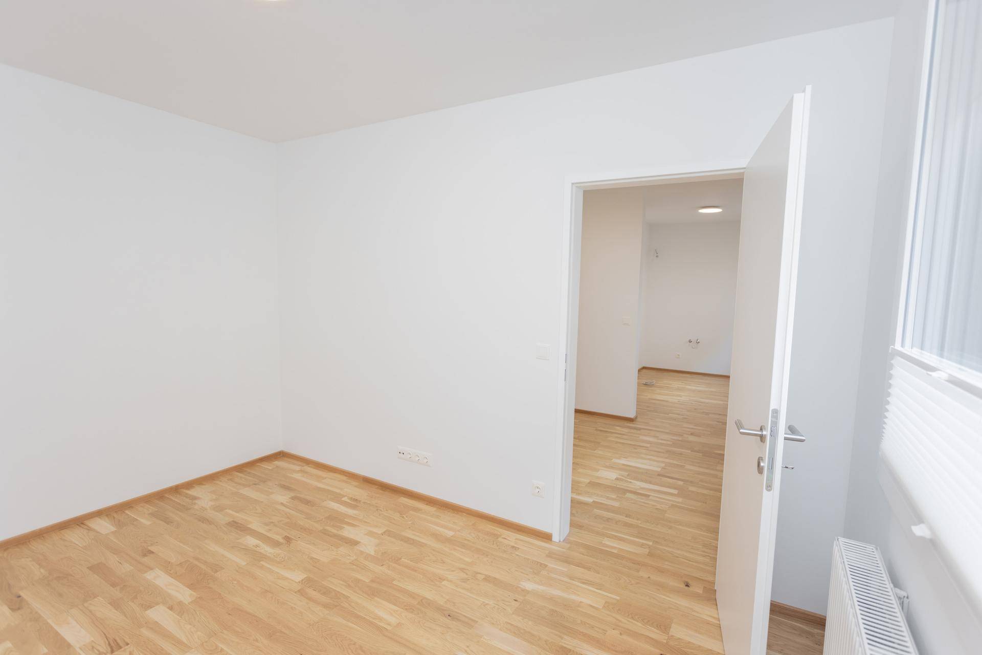 Leerer Raum mit Parkettboden und weißen Wänden, der den Blick in angrenzende Bereiche ermöglicht.