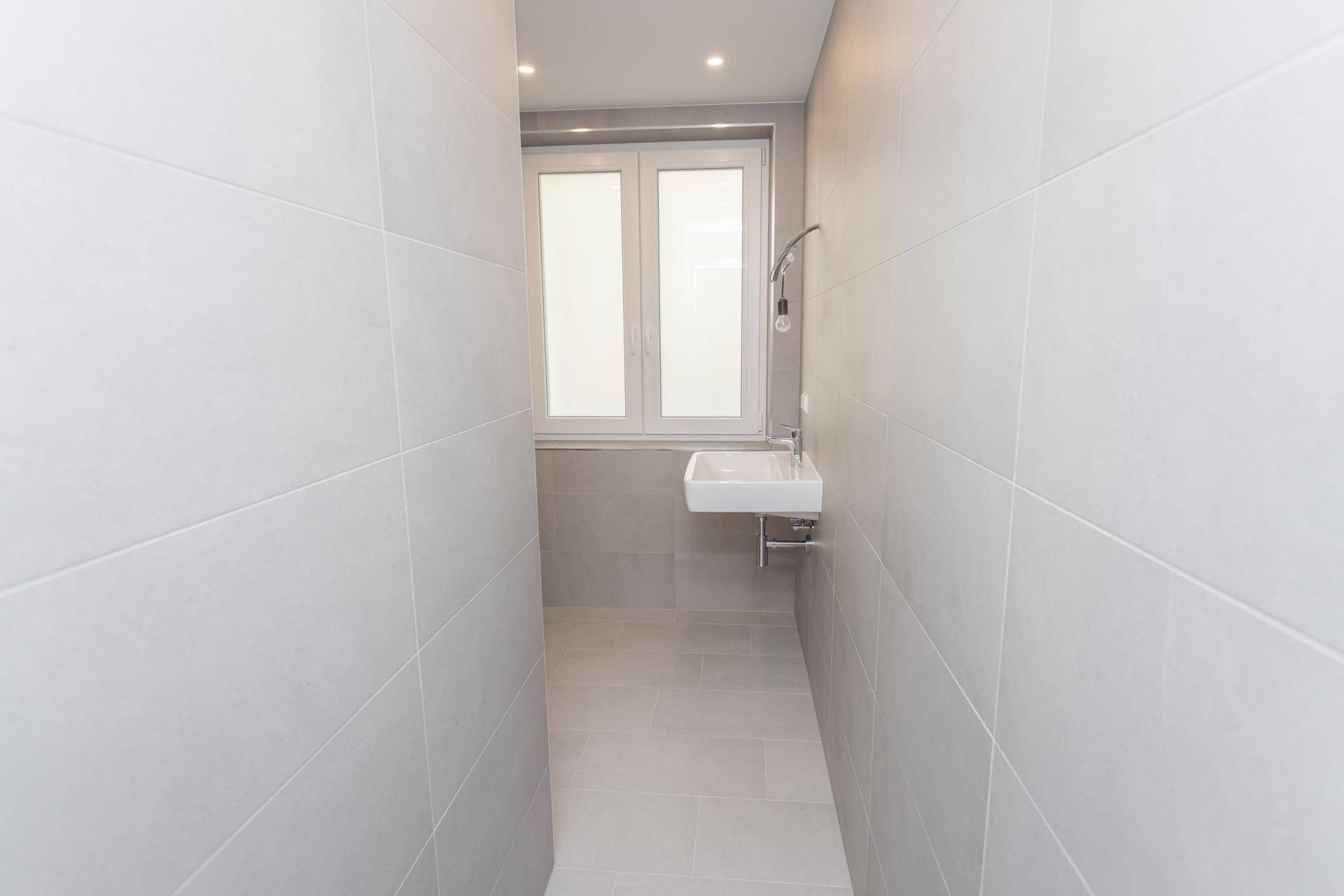 Schmaler, heller Badezimmerbereich mit Waschbecken, Fenster und grauen Wandfliesen.