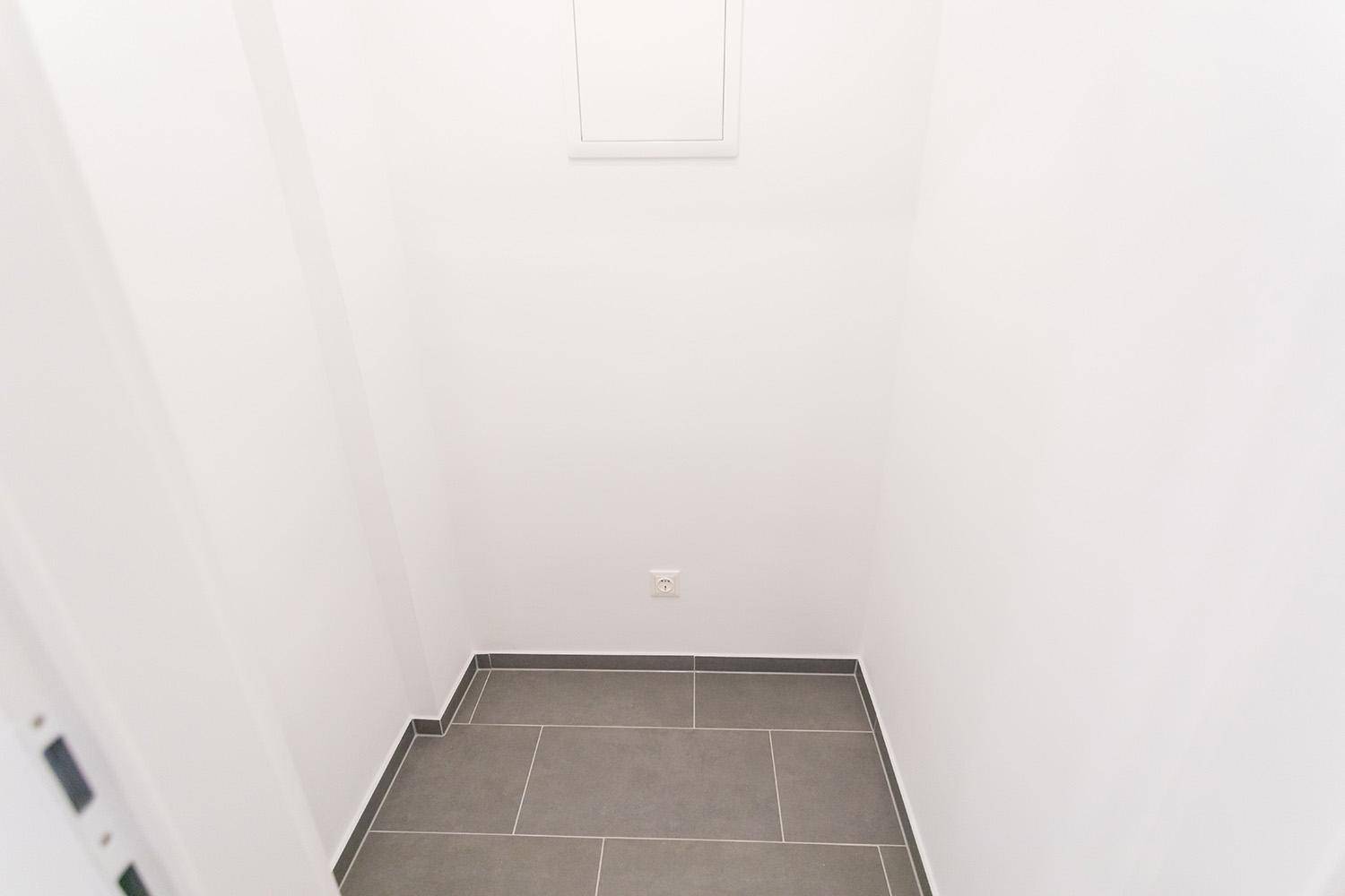 Leerer Abstellraum mit weißen Wänden und grauen Bodenfliesen.