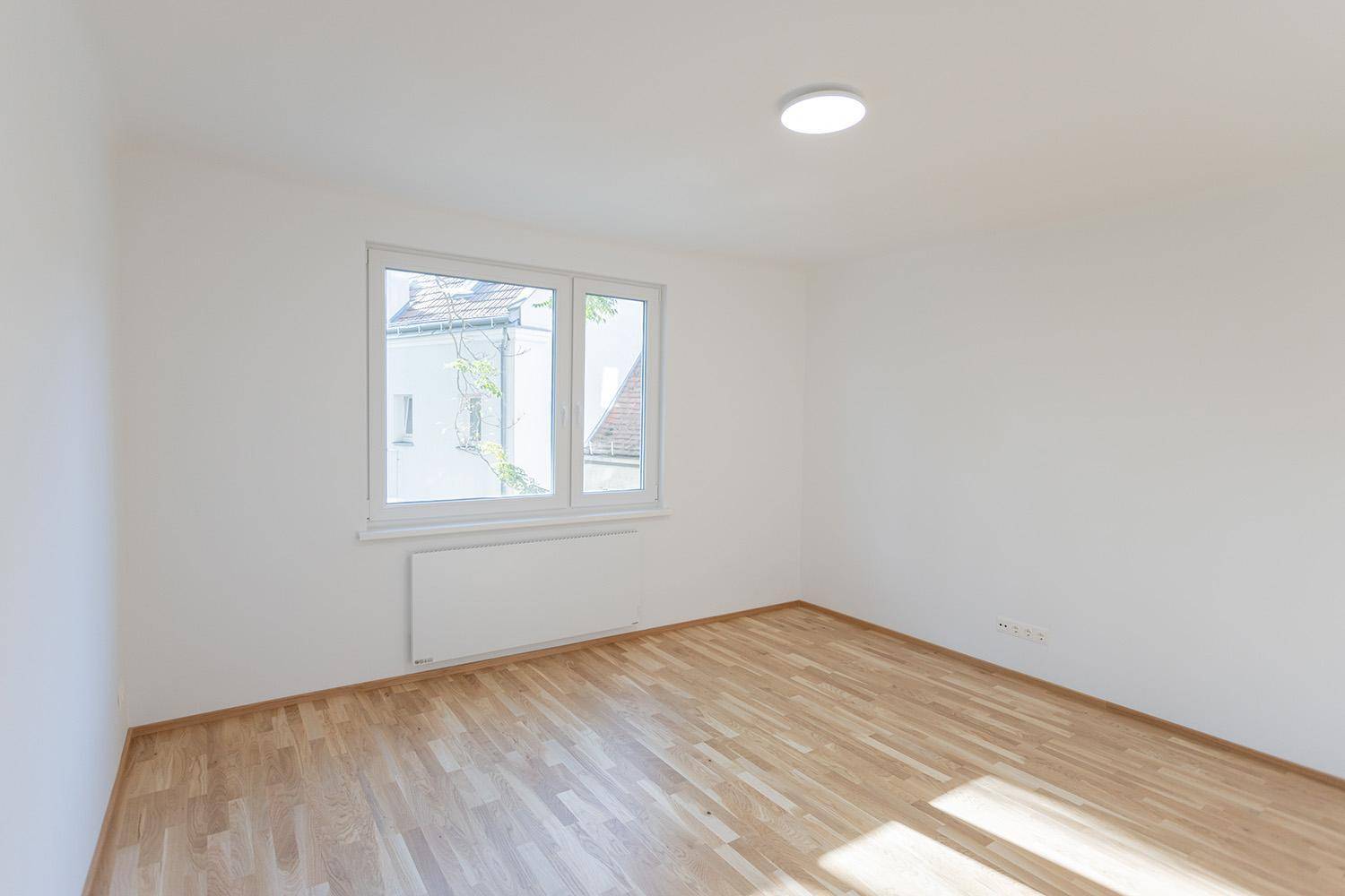 Leerer, heller Raum mit Holzboden und Fenster, das einen Blick ins Grüne bietet.