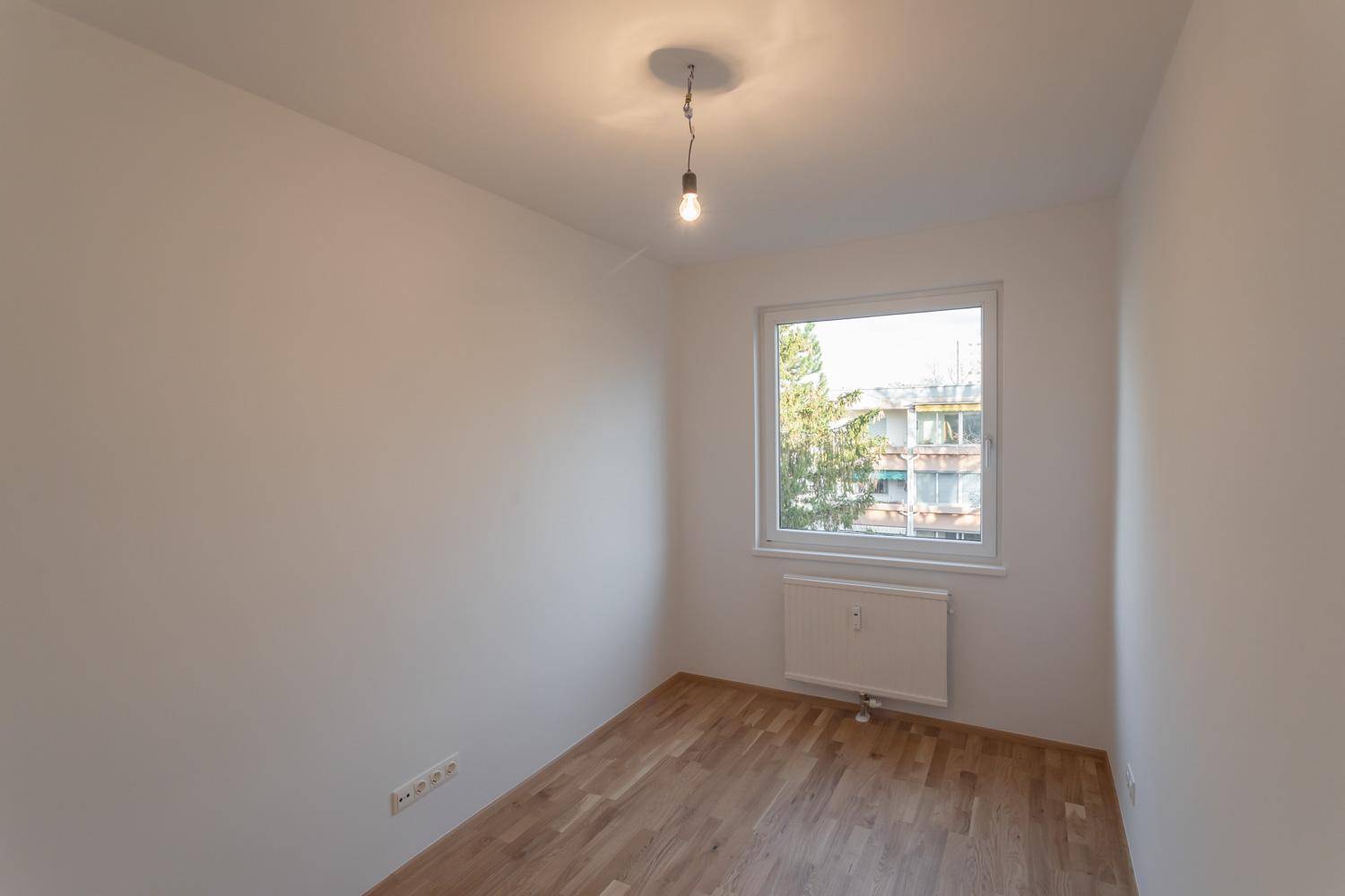 Leeres Zimmer mit Parkettboden und Fenster, das viel Tageslicht hereinlässt.