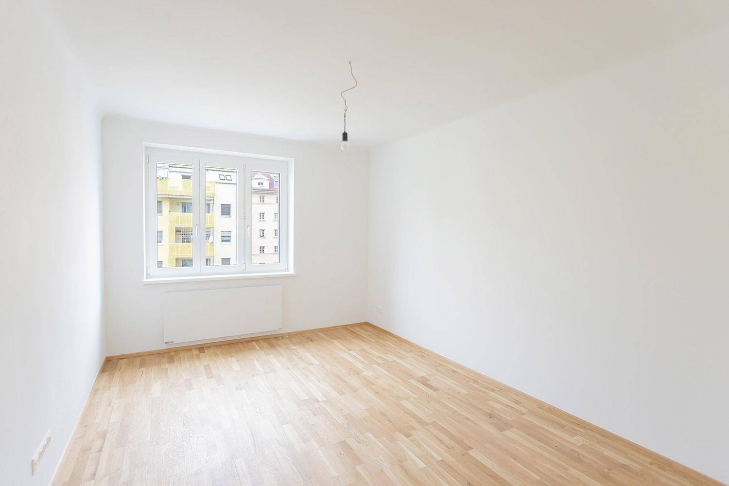 Geräumiges, leeres Zimmer mit Parkettboden und einem Fenster mit Blick auf ein Mehrfamilienhaus.