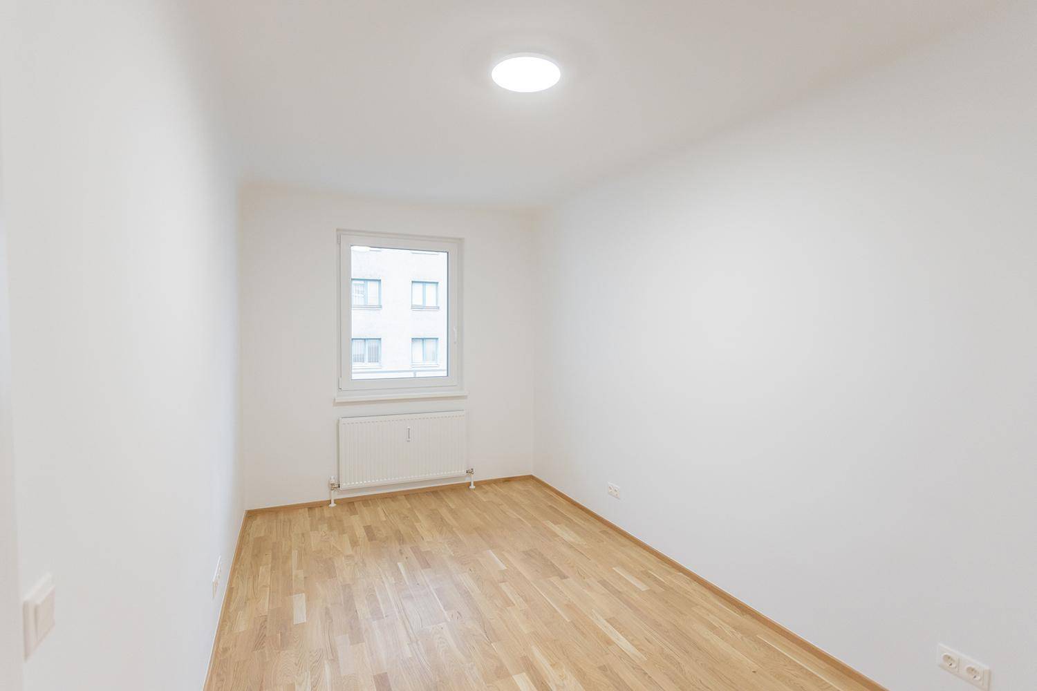 Helles, leeres Zimmer mit Parkettboden und einem Fenster, das auf ein Gebäude blickt.
