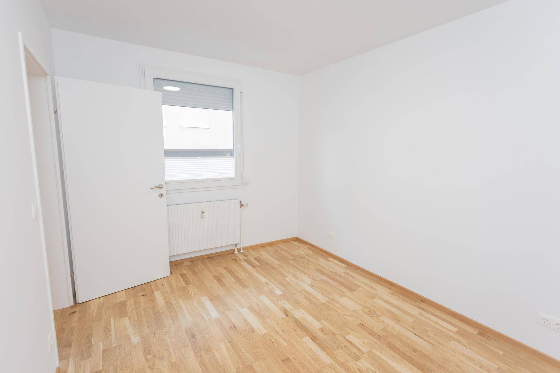 Unmöblierter Raum mit Holzboden, weißen Wänden und einem Fenster mit Rollladen für Privatsphäre.