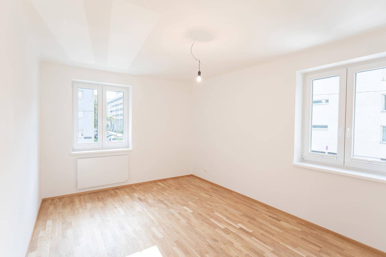 Helles Zimmer mit zwei Fenstern, Parkettboden und weiß gestrichenen Wänden.