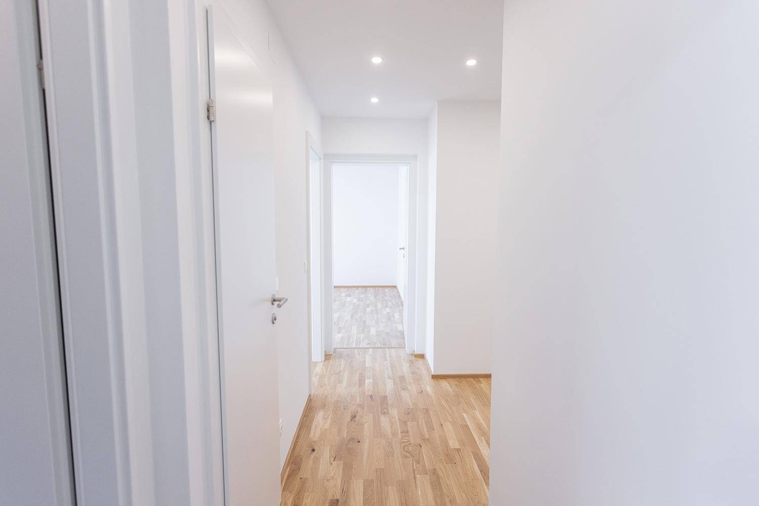Heller Flur mit Holzparkettboden und weißen Wänden, der einen Blick in angrenzende Räume bietet.