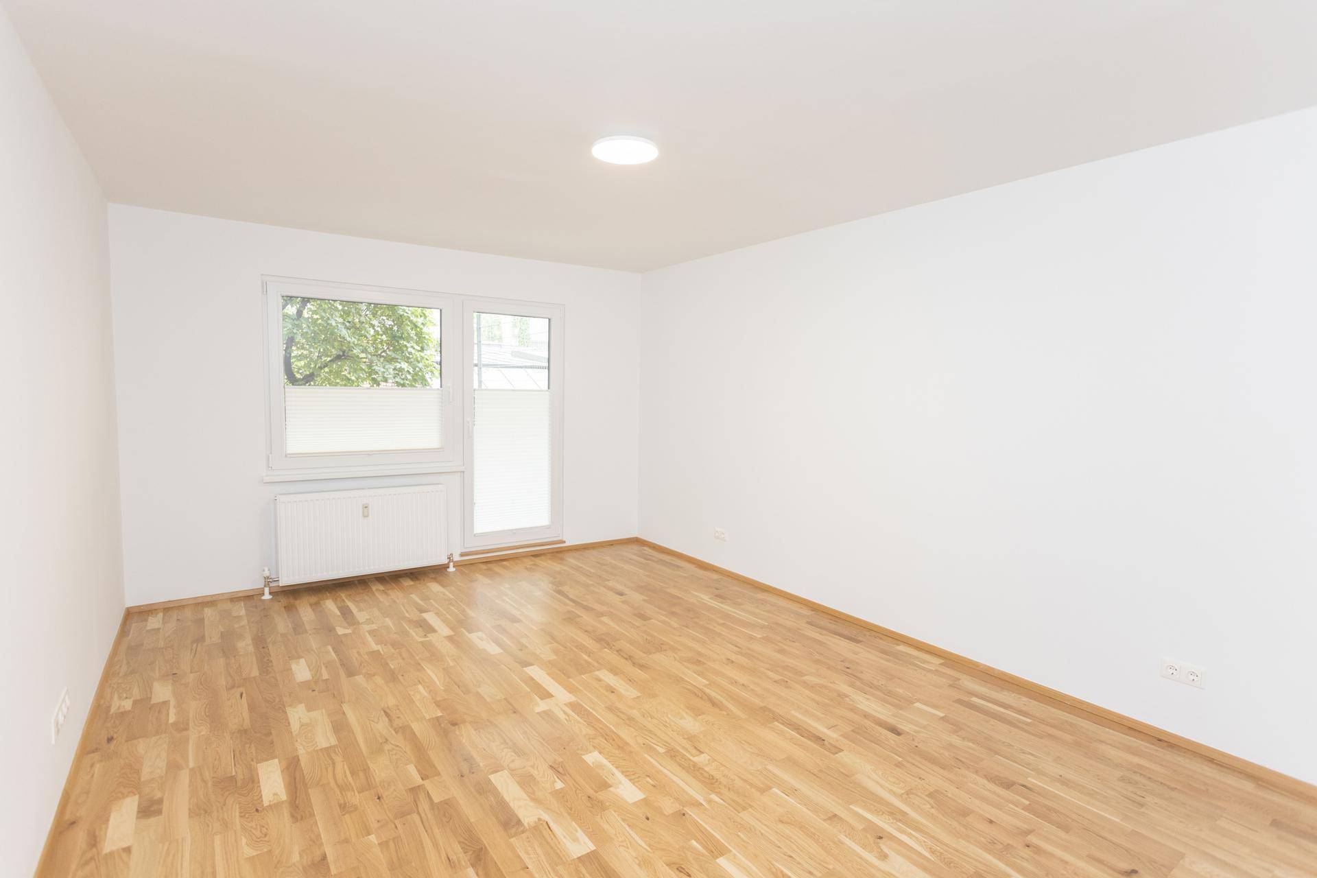 Heller, leerer Raum mit weiß gestrichenen Wänden, Holzparkettboden und großem Fenster.