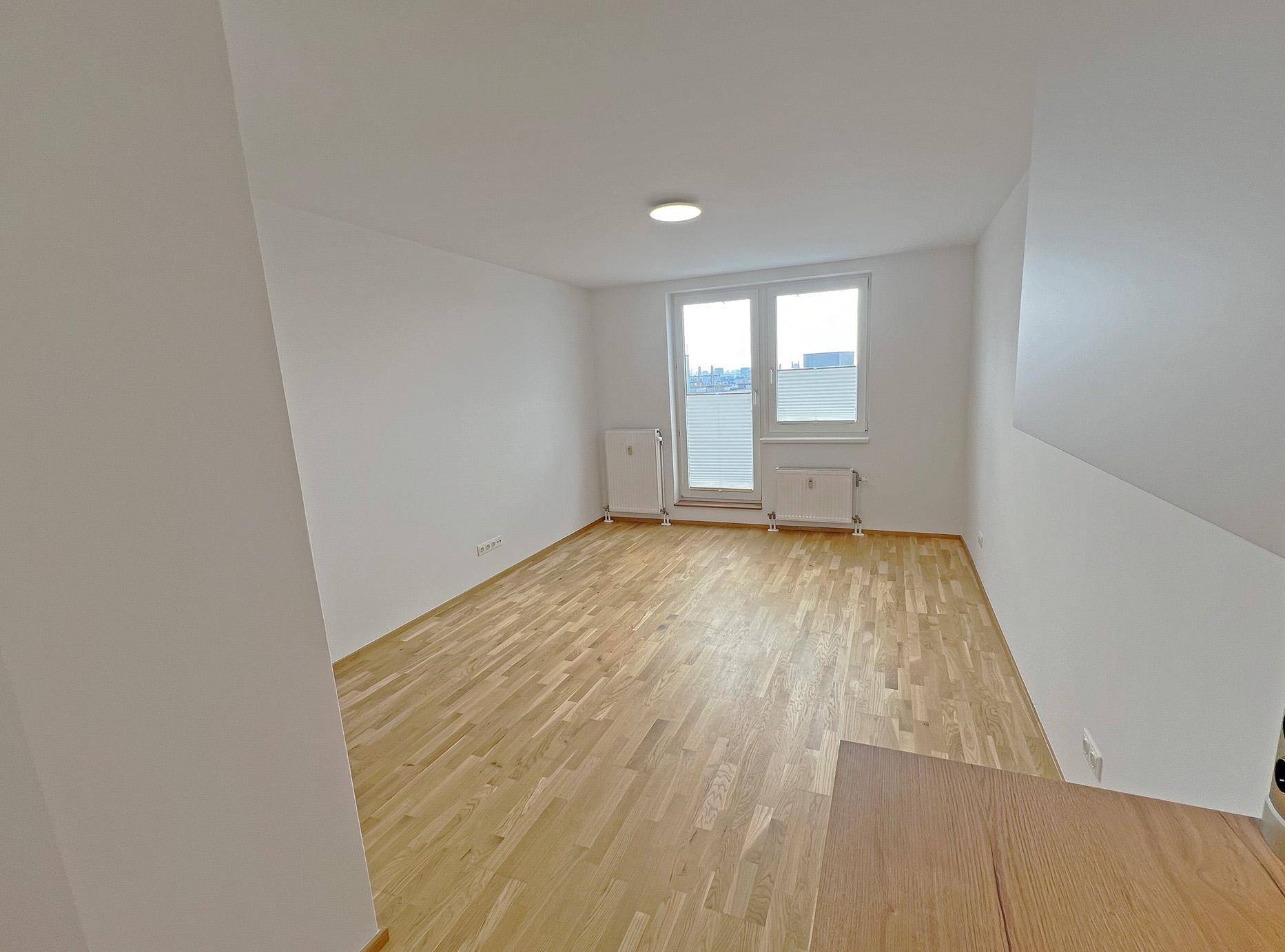 Helles, leeres Zimmer mit Parkettboden und Fenster mit Jalousien und Stadtblick.