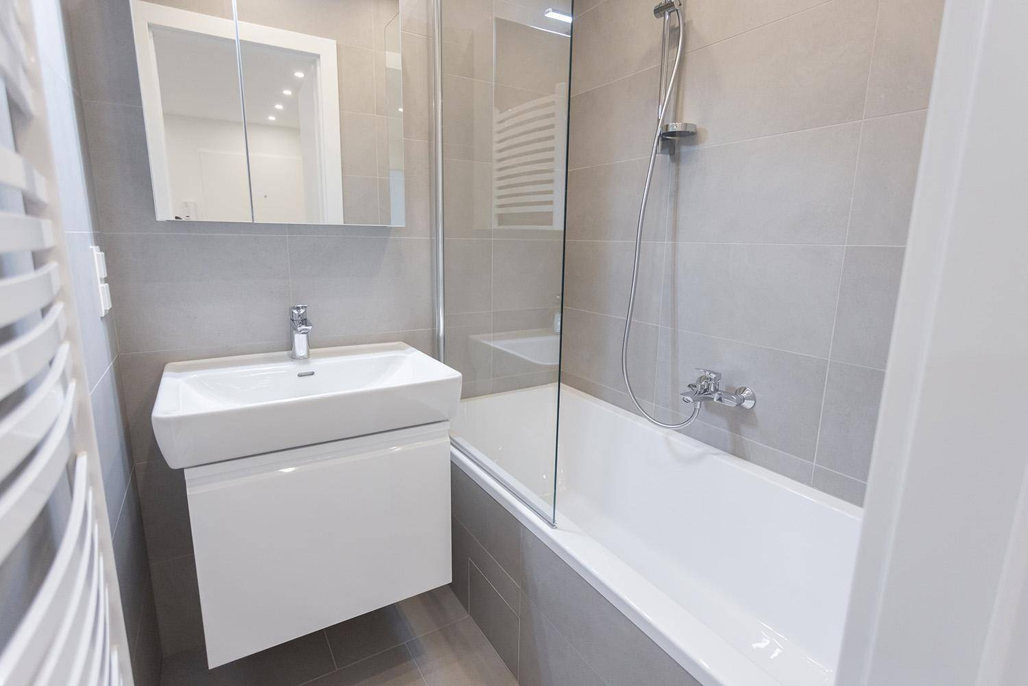 Modernes Badezimmer mit grauen Fliesen, Waschtisch und Badewanne mit Duschabtrennung aus Glas.