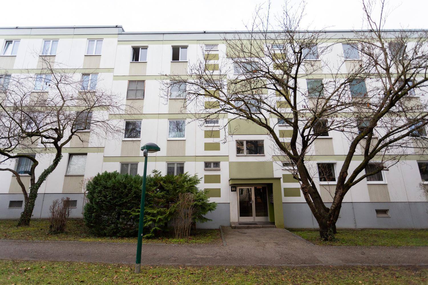 Mehrfamilienhaus mit grüner Fassadengestaltung und Bäumen im Vordergrund an einem bewölkten Tag.