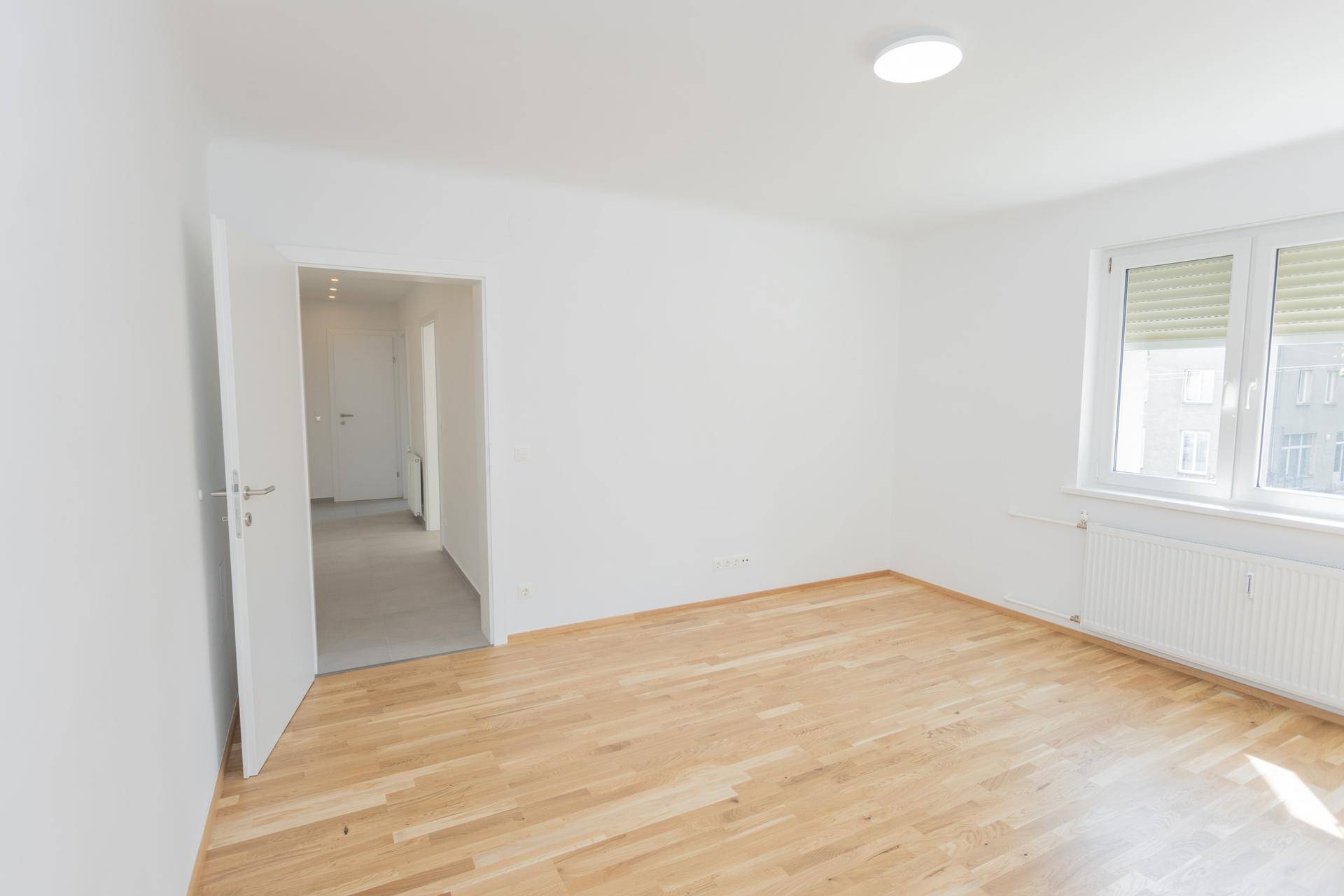 Leeres, helles Schlafzimmer mit Holzboden und offener Tür zum Flur, viel Tageslicht.
