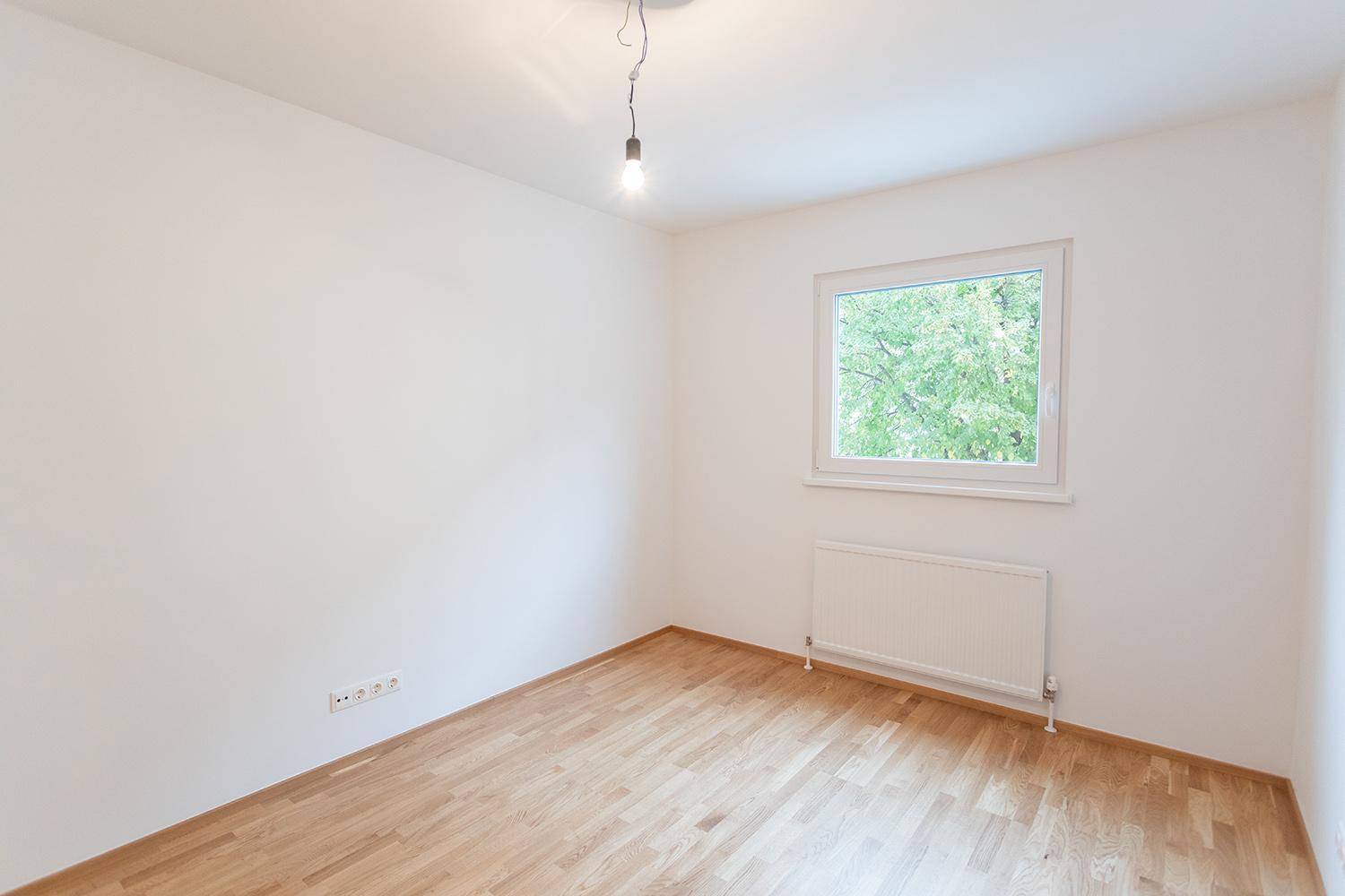 Leeres Schlafzimmer mit hellem Holzboden, weiß gestrichenen Wänden und Fenster mit Ausblick.