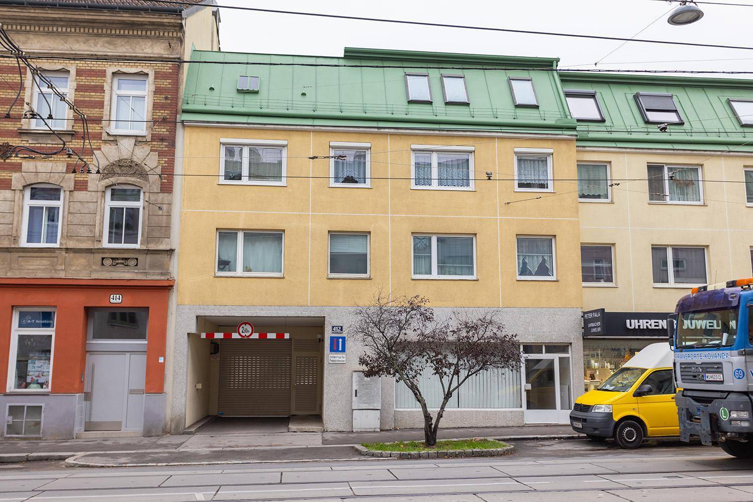 Mehrfamilienhaus mit gelber Fassade, grünem Dach und Garageneinfahrt an einer Stadtstraße.