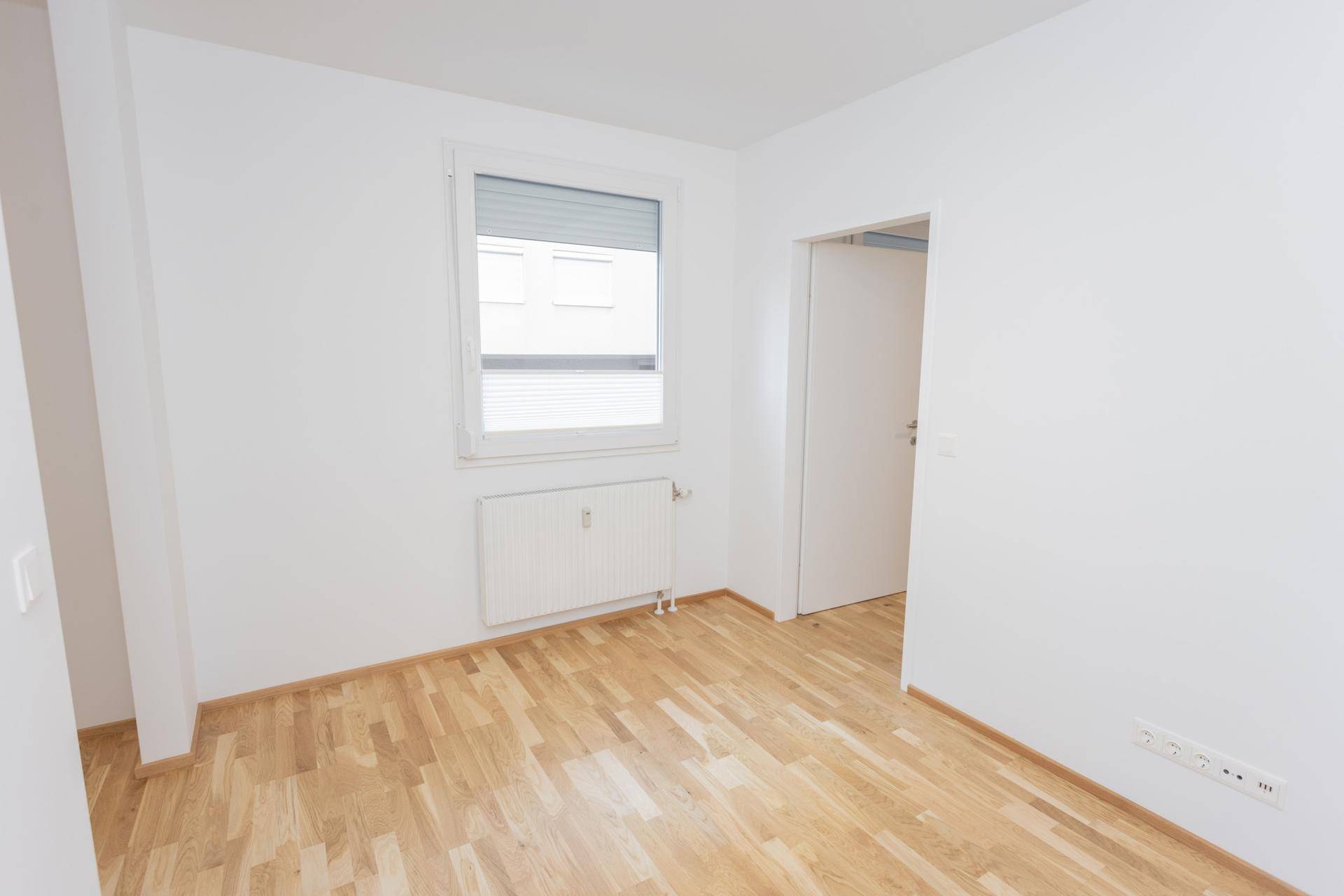 Heller, leerer Raum mit Parkettboden, Fenster mit Rollläden und Heizkörper, bereit zur Einrichtung.