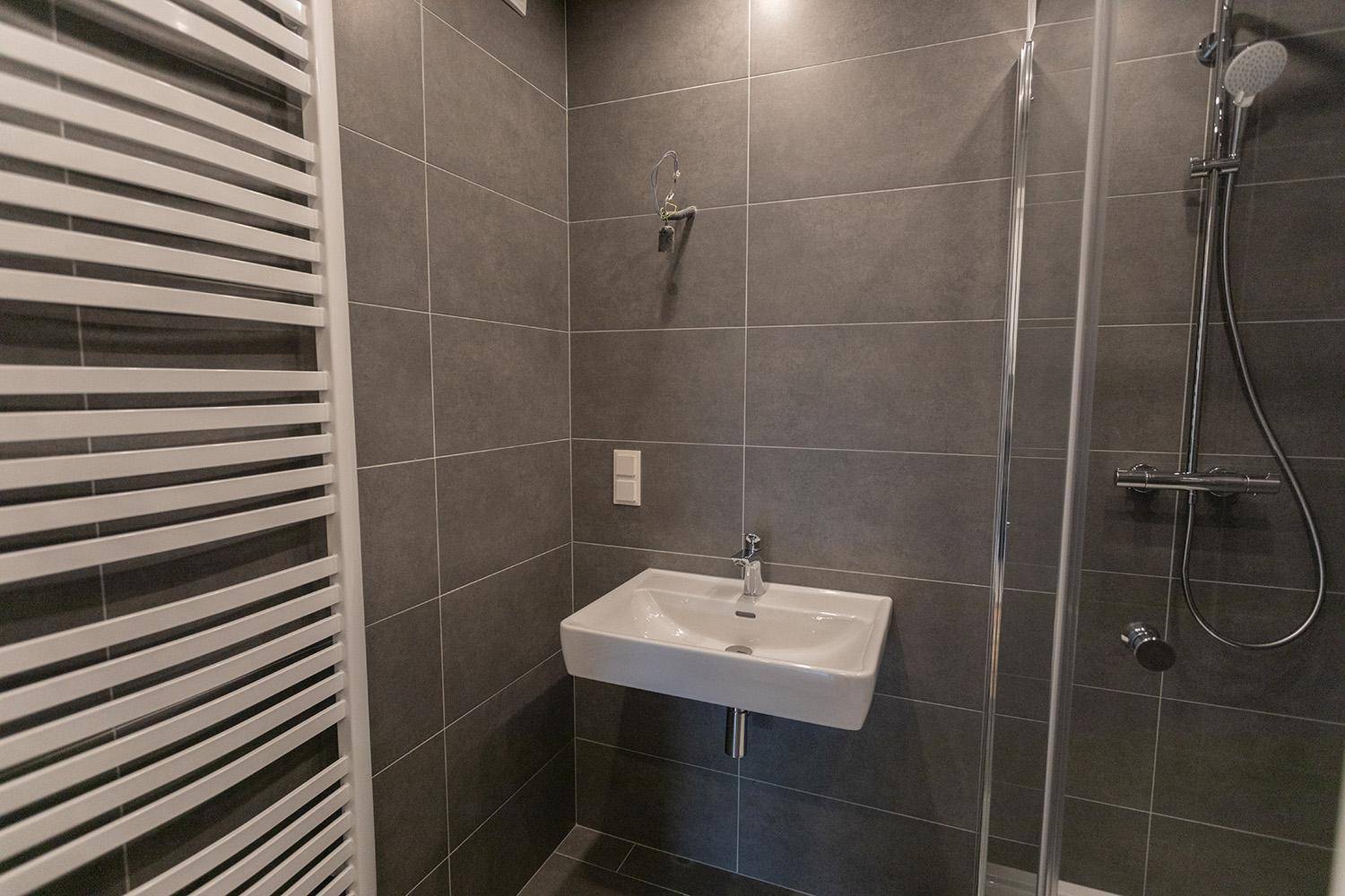 Modernes Badezimmer mit grauen Fliesen, Duschkabine und wandhängendem Waschbecken.
