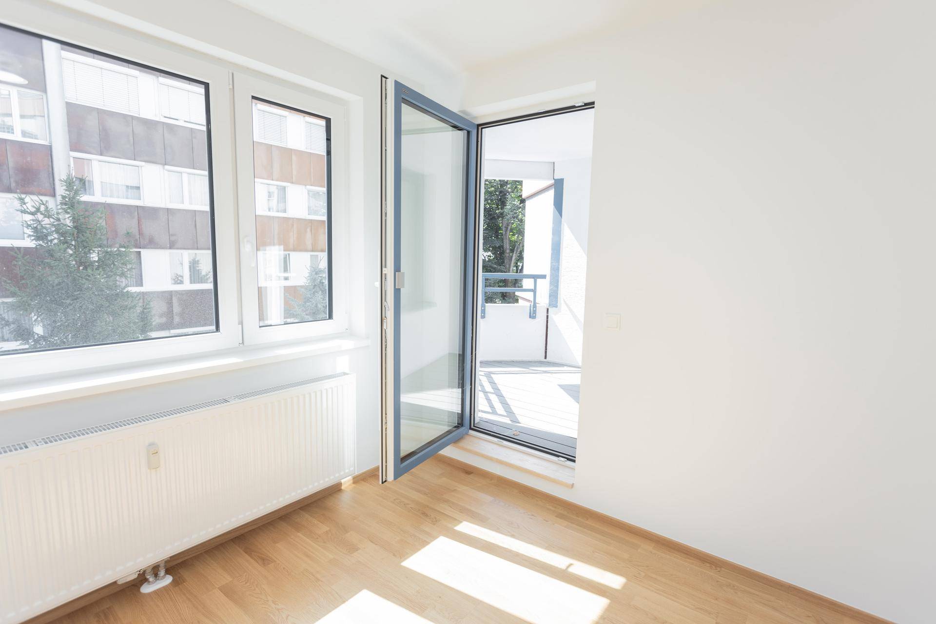 Lichtdurchflutetes Zimmer mit Holzparkett und direktem Zugang zu einem Balkon, mit Blick auf ein Wohngebäude.