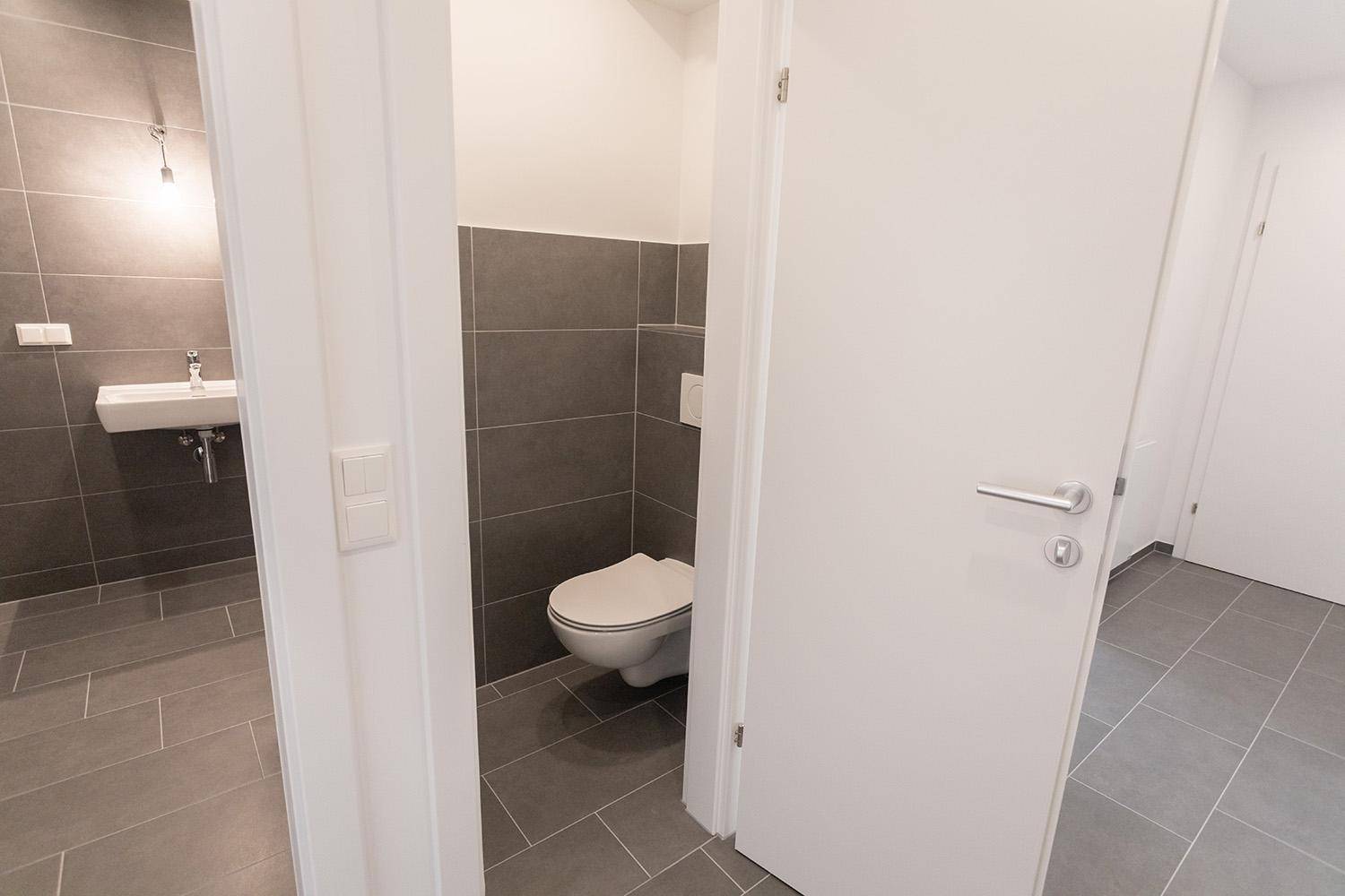 Separate Toilette mit Wand-WC, kleinem Waschbecken und modernen grauen Fliesen, praktisch und sauber.