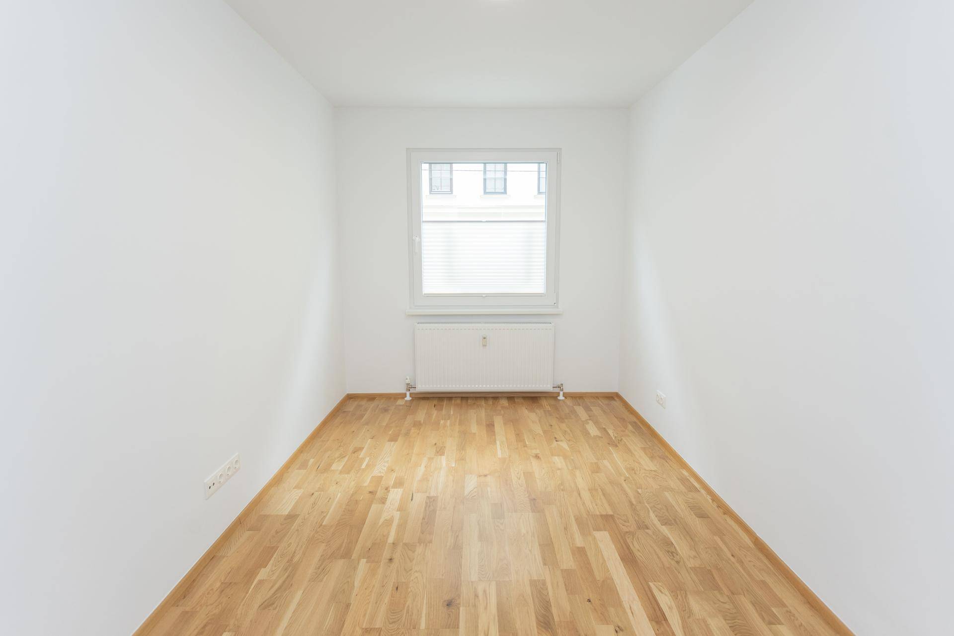 Kleines, helles Zimmer mit Parkettboden und einem Fenster mit Blick nach draußen.