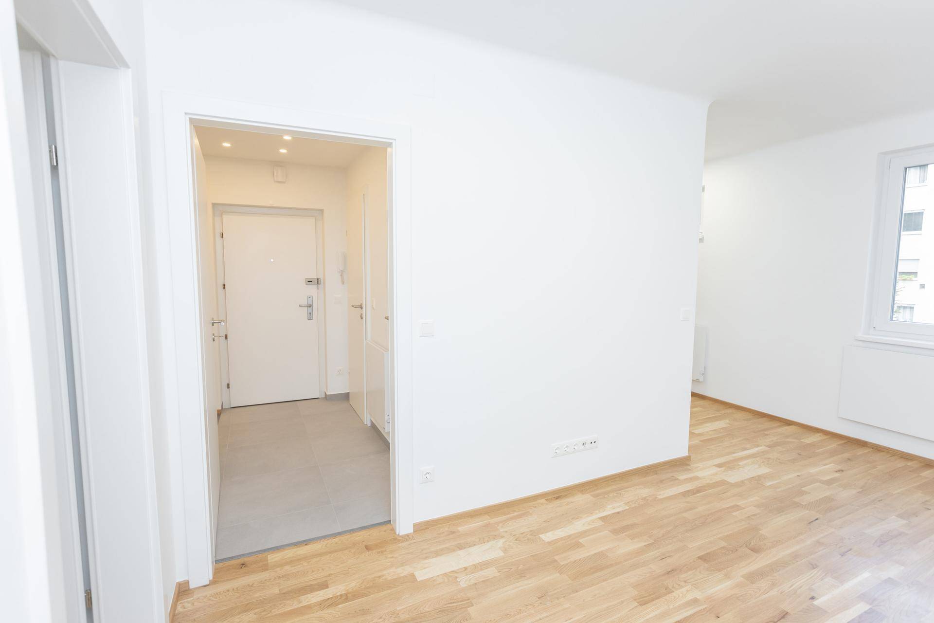 Heller, leerer Raum mit Holzfußboden, der zum Eingangsbereich mit Fliesenboden führt.