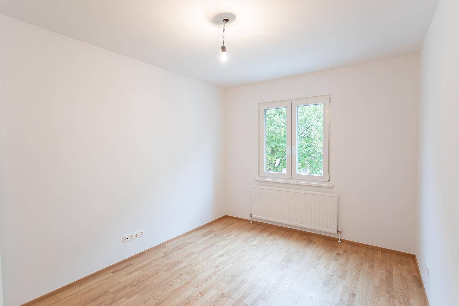 Leeres Schlafzimmer mit hellem Holzboden, weiß gestrichenen Wänden und großem Fenster.