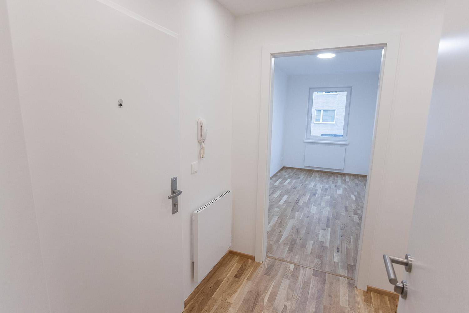 Heller Flur mit Holzboden und offener Tür zu einem leeren Zimmer mit Fenster.